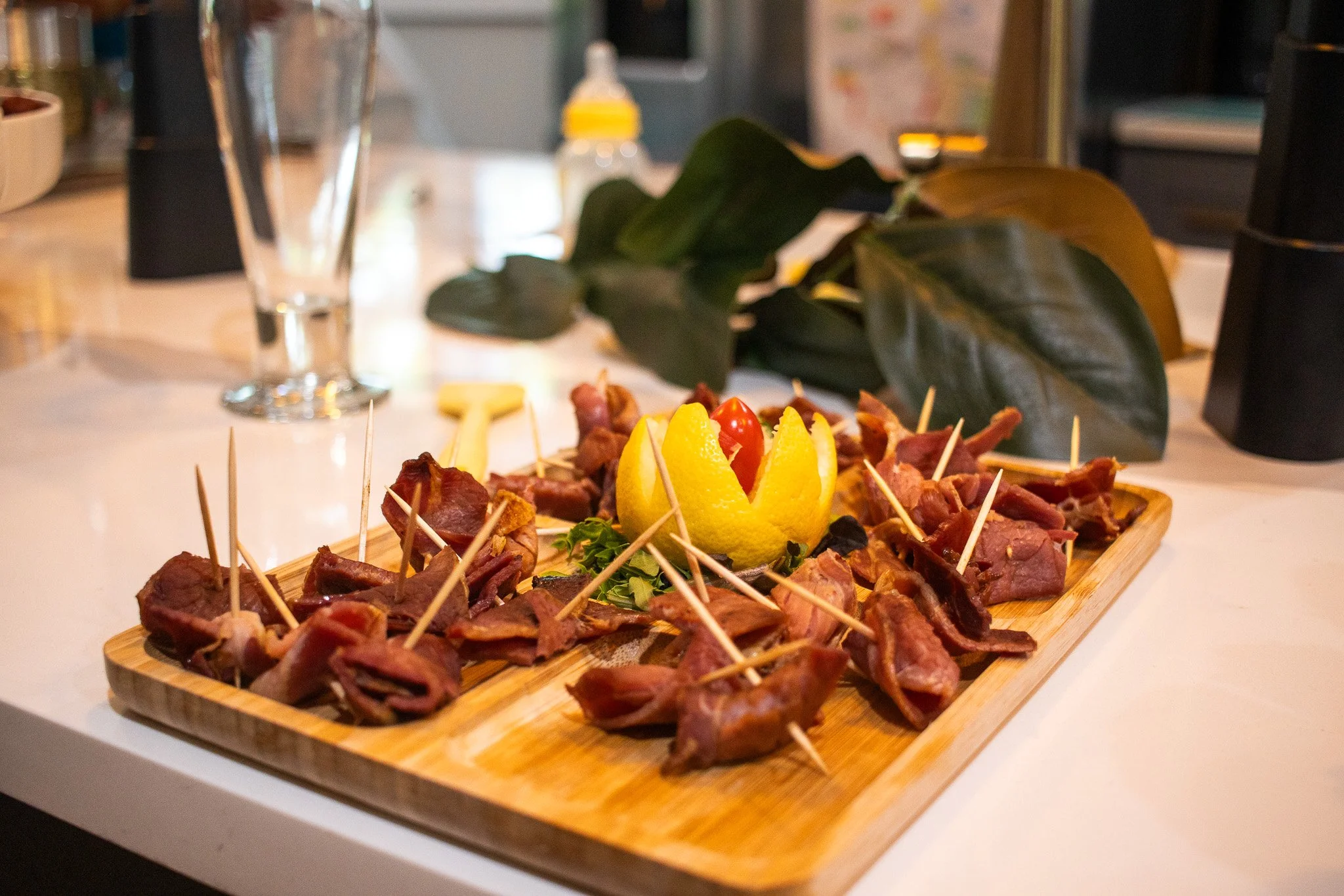 A wooden platter with bacon-wrapped appetizers, garnished with lemon slices and cherry tomatoes, set on a white table with a glass of water and a baby bottle in the background.