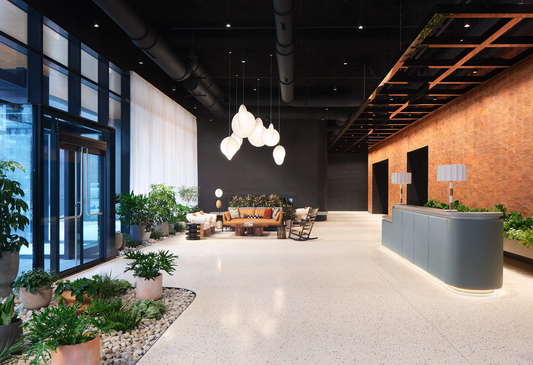 Modern lobby with large glass doors, potted plants, a seating area with sofas and chairs, hanging pendant lights, and a reception desk with lamps, with a brick wall and black ceiling with exposed ducts.