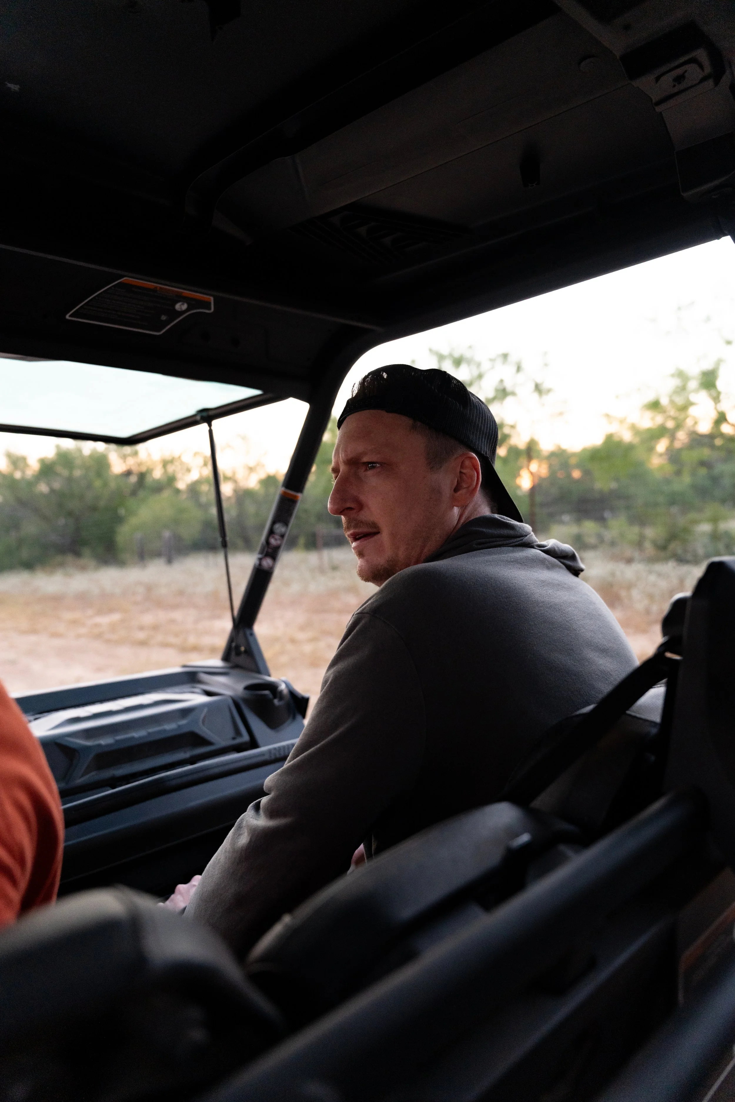 A man with a backward cap in a vehicle, looking to the side with a focused expression, with open landscape and trees in the background.