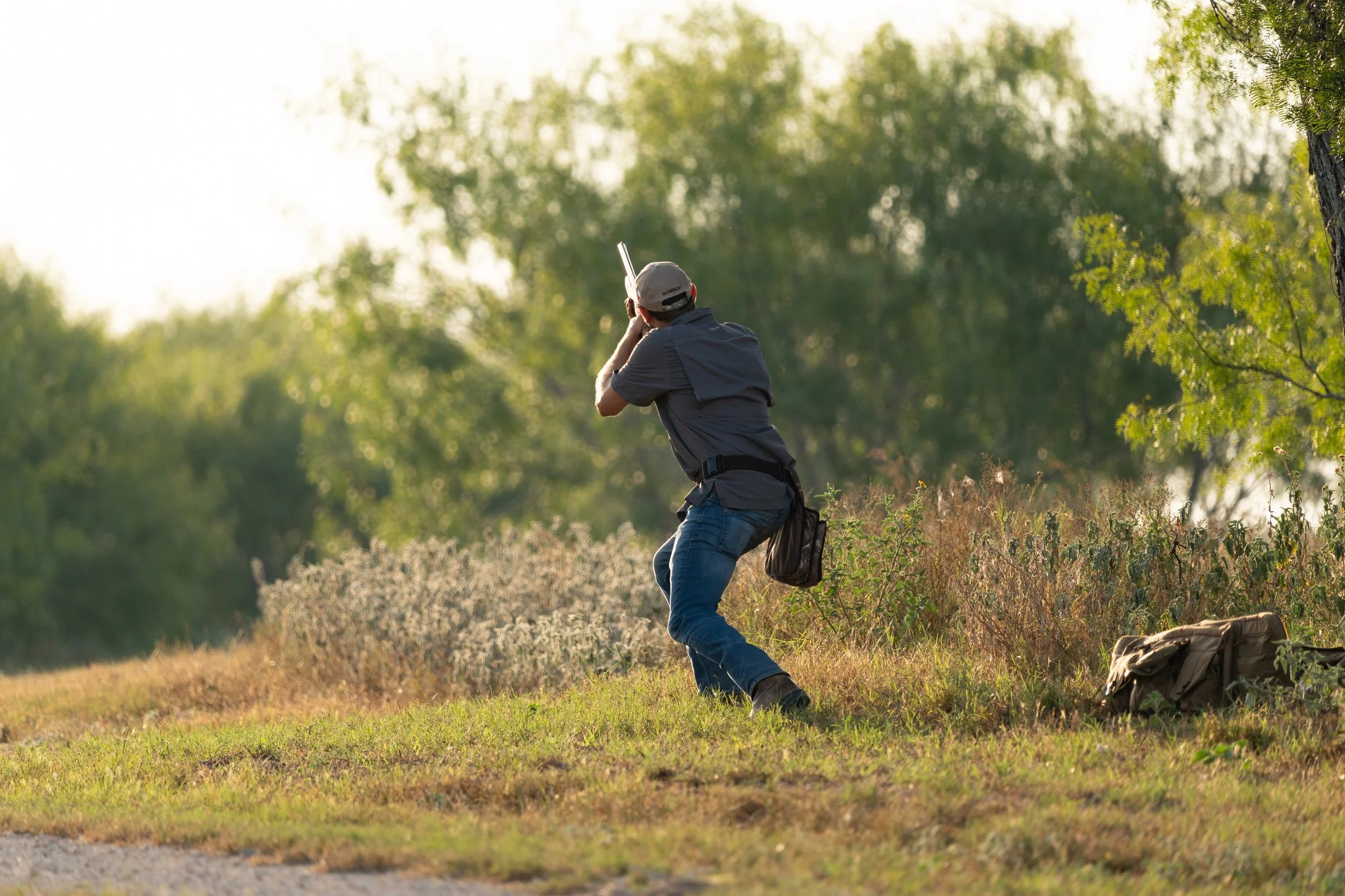 A person aiming a shotgun outdoors in a grassy area with trees in the background.