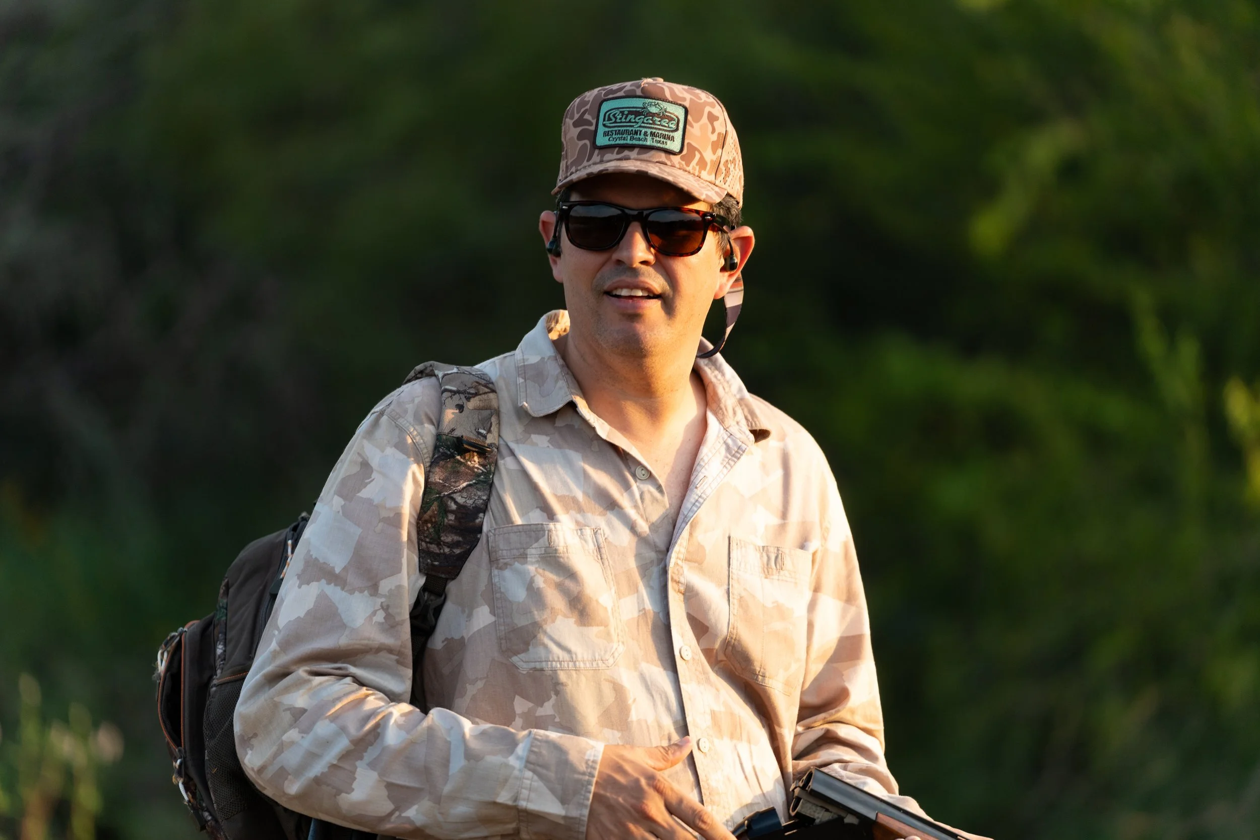 A man wearing a camouflage baseball cap, sunglasses, and a beige camouflage shirt outdoors. He carries a backpack and holds a device in his hand. Green foliage is in the background.