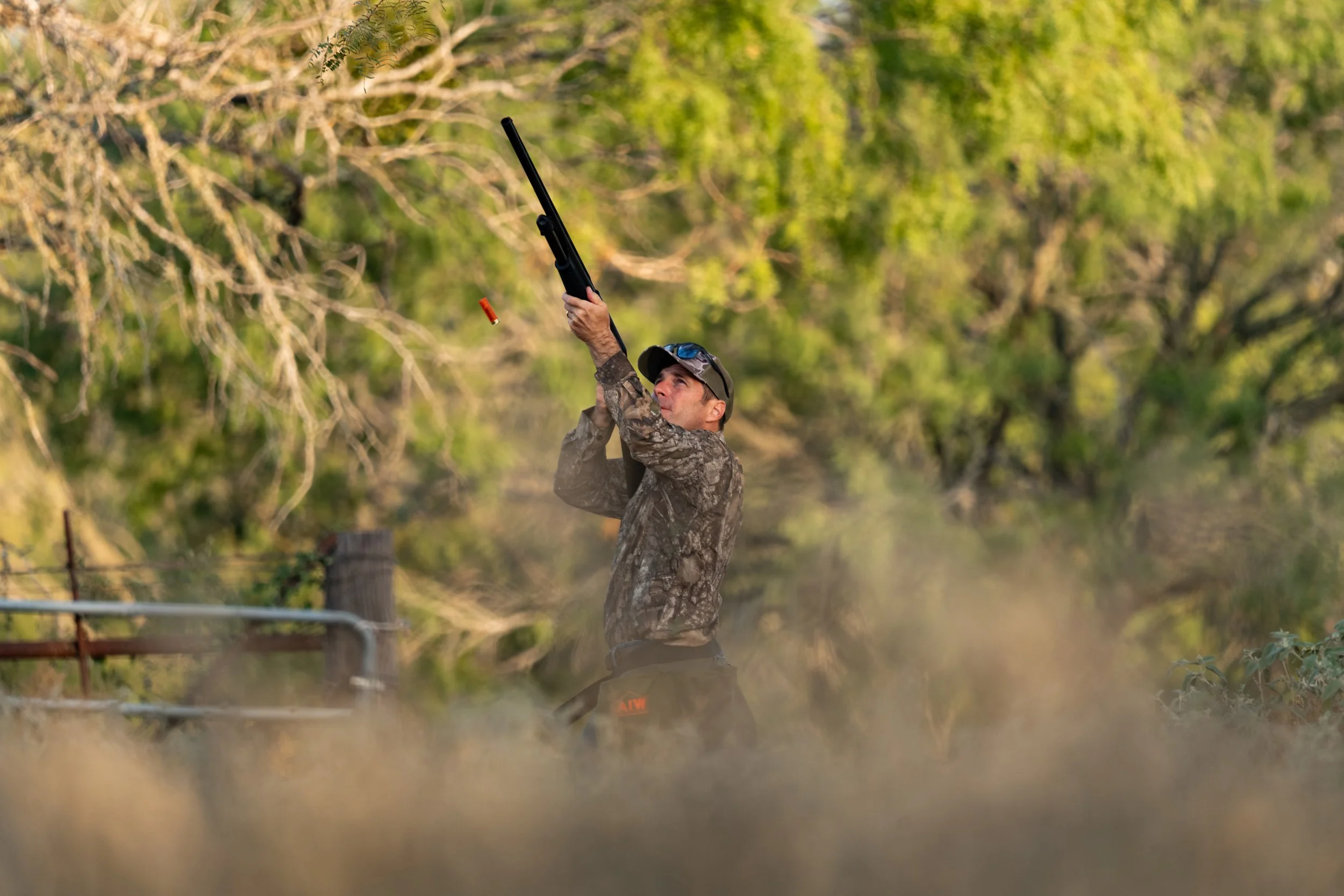 A man dressed in camouflage hunting gear aiming a shotgun upward, with a clay target flying in the air, outdoors in a natural setting with green trees and grass.