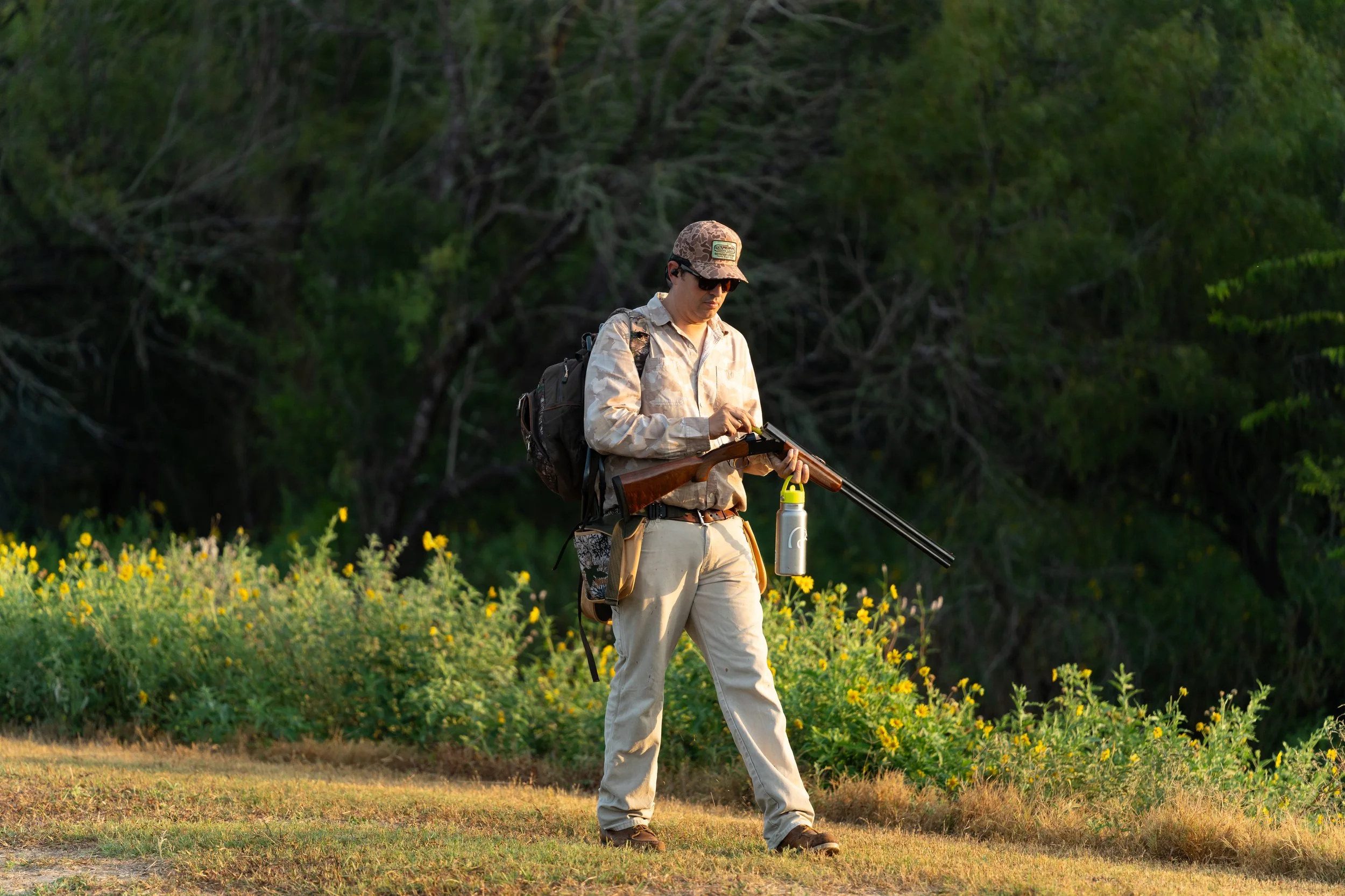 A man in outdoor clothing with a backpack, wearing sunglasses and a cap, is holding a rifle with one hand and adjusting it with the other in a natural, green setting.