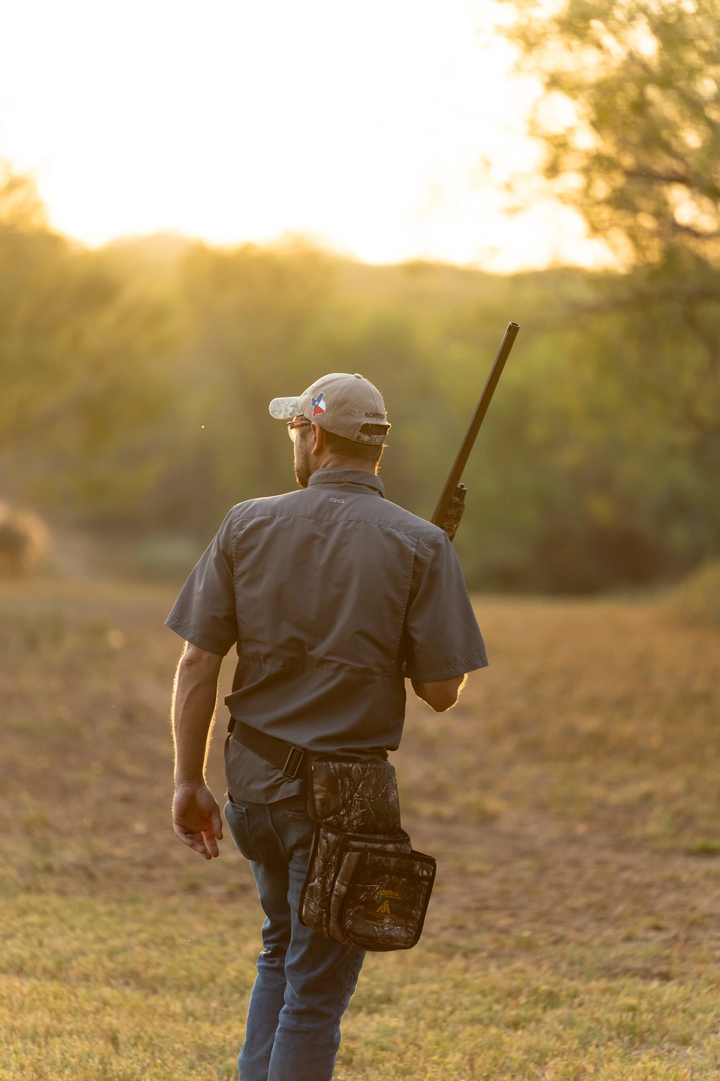 A man with a hat and sunglasses walking outdoors holding a shotgun over his shoulder, with trees and sunlight in the background.