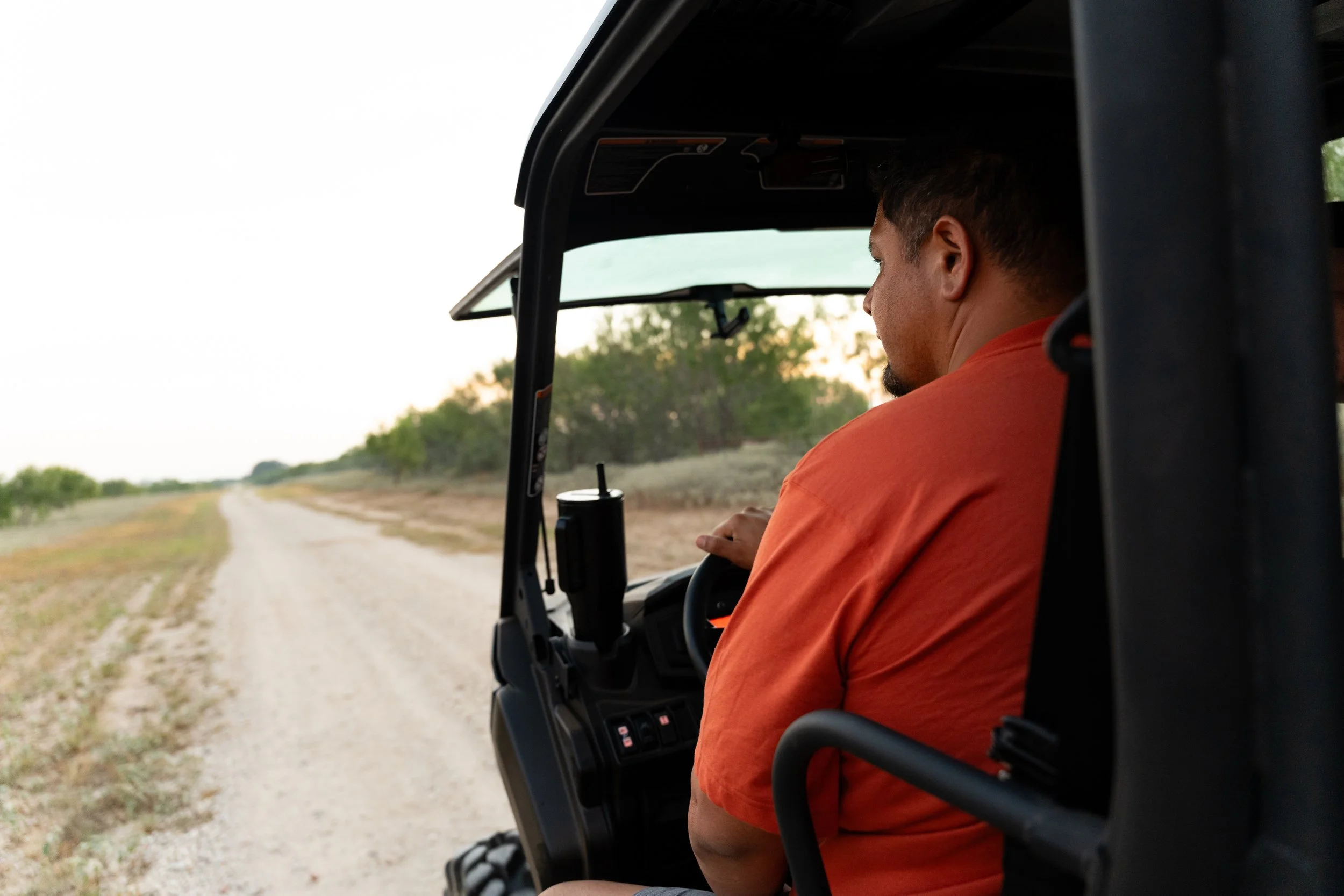 Man driving an ATV or utility vehicle on a dirt trail surrounded by trees at sunset.