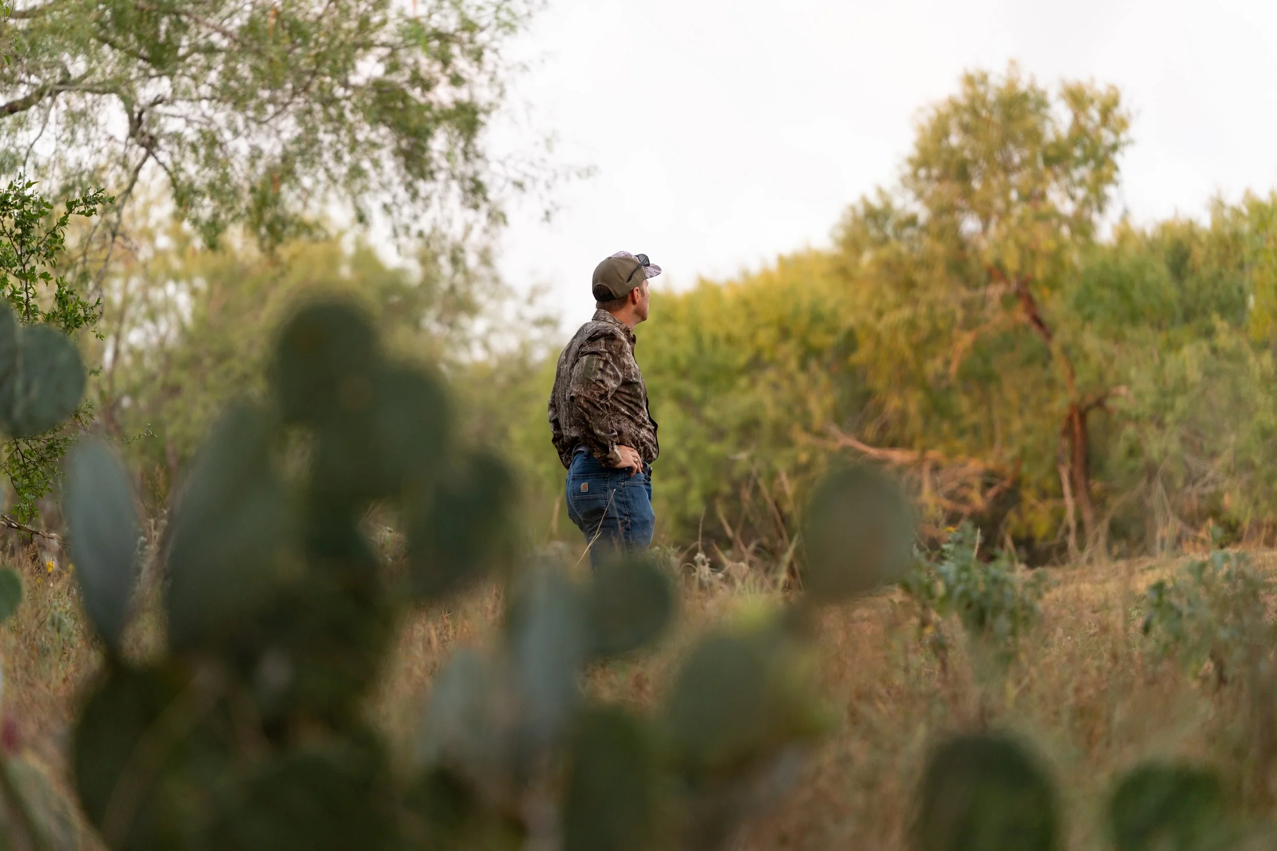A man in camouflage clothing standing outdoors among greenery and bushes, looking away into the distance during daytime.