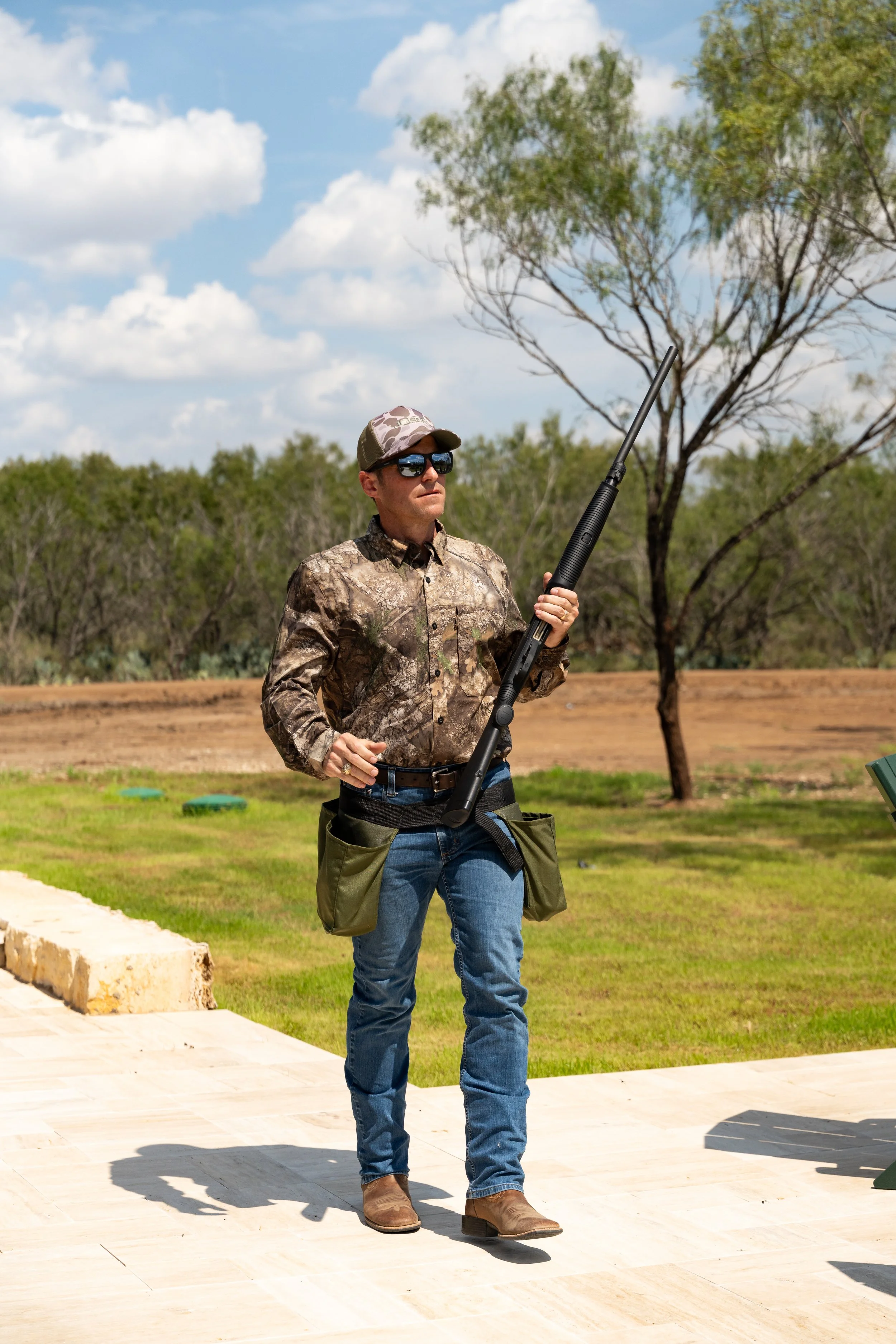 A person in camo clothing holding a rifle at an outdoor shooting range with grass, trees, and a blue sky with clouds in the background.