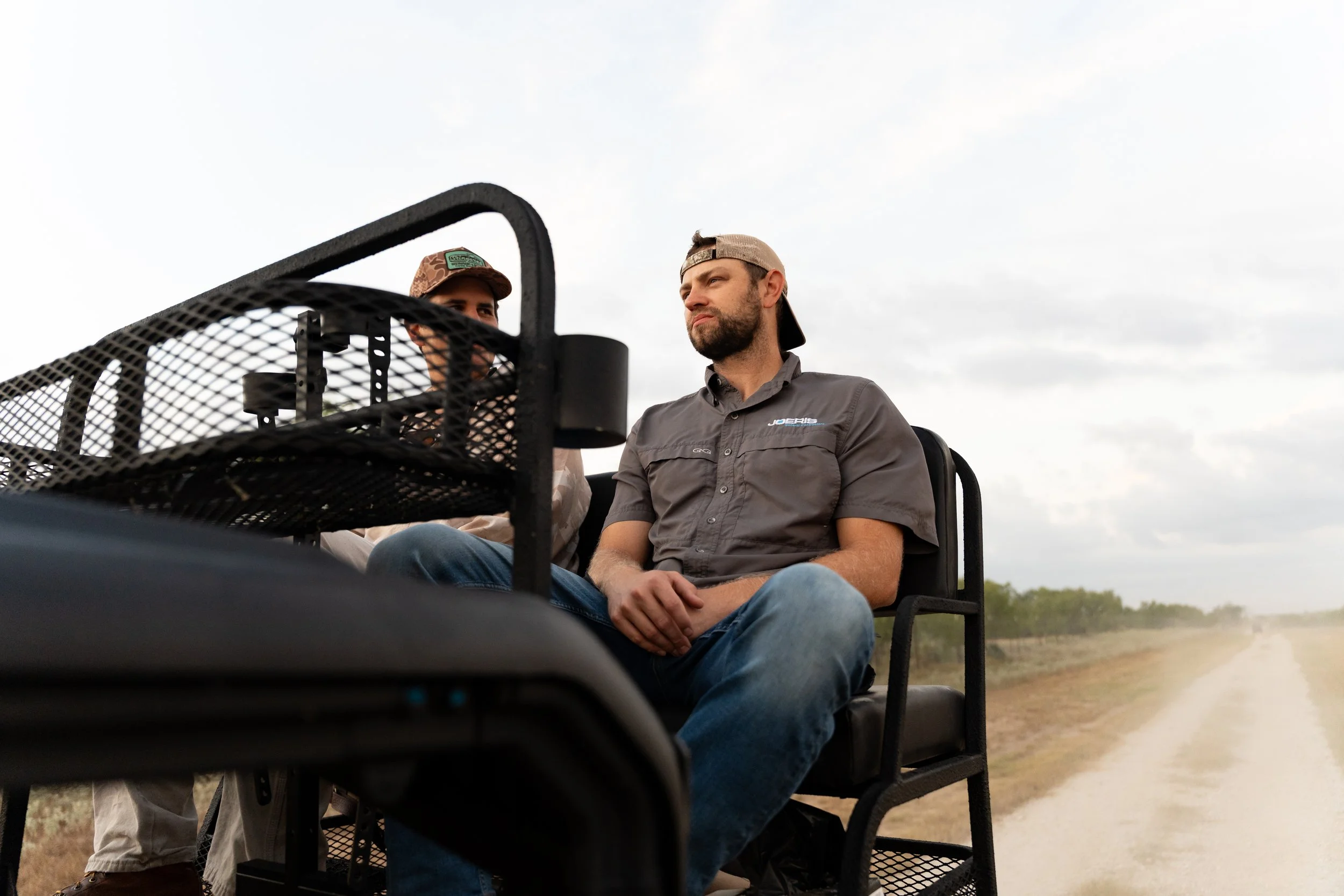 Two men sit on a vehicle in a rural area, one wearing a dark gray shirt and backwards cap, the other in a beige shirt and cap, with a dirt road and open landscape in the background.