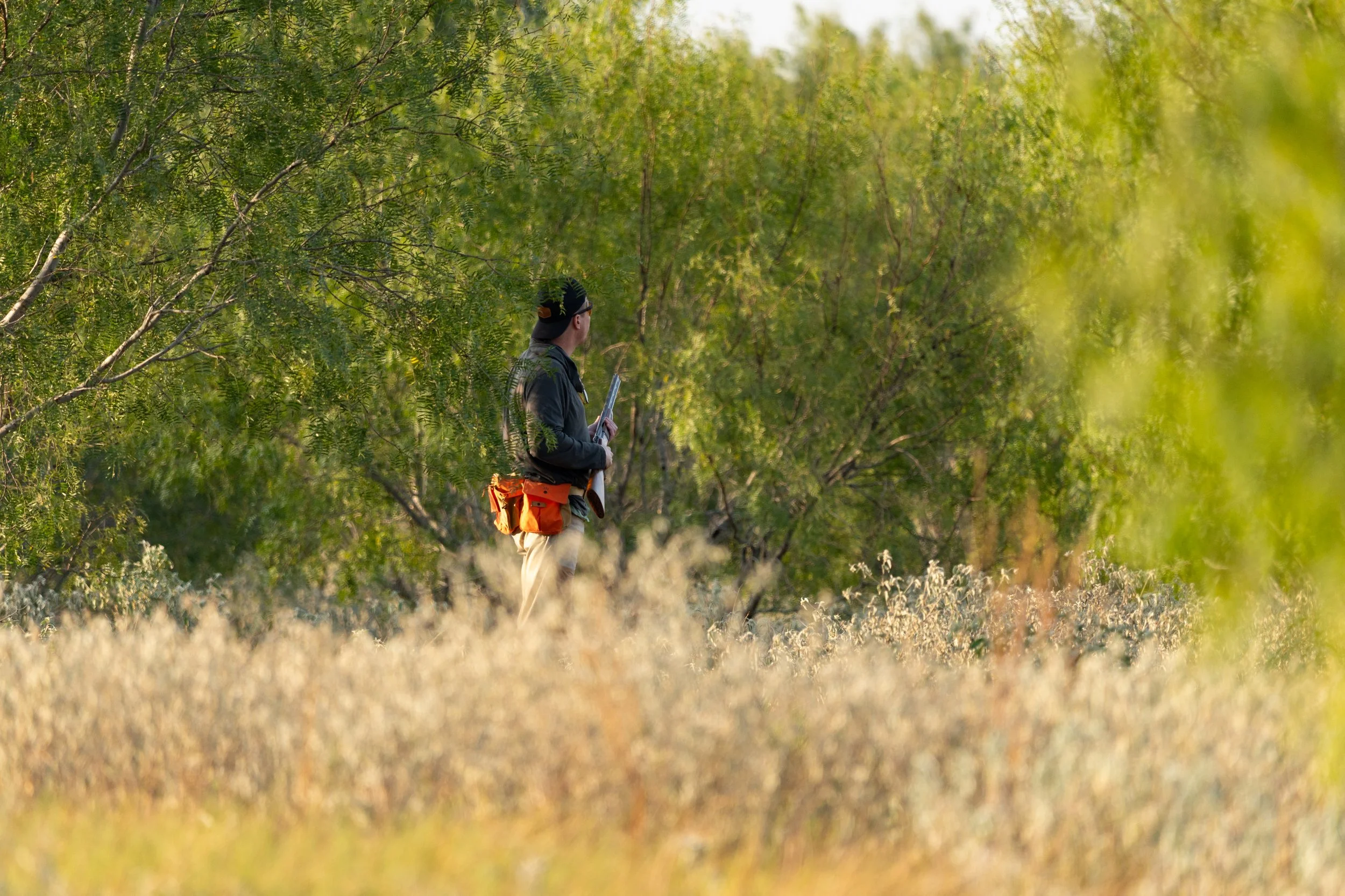 A person standing among green trees and tall grass, wearing a dark jacket, light-colored pants, a black cap, and carrying an orange backpack while holding a clipboard.