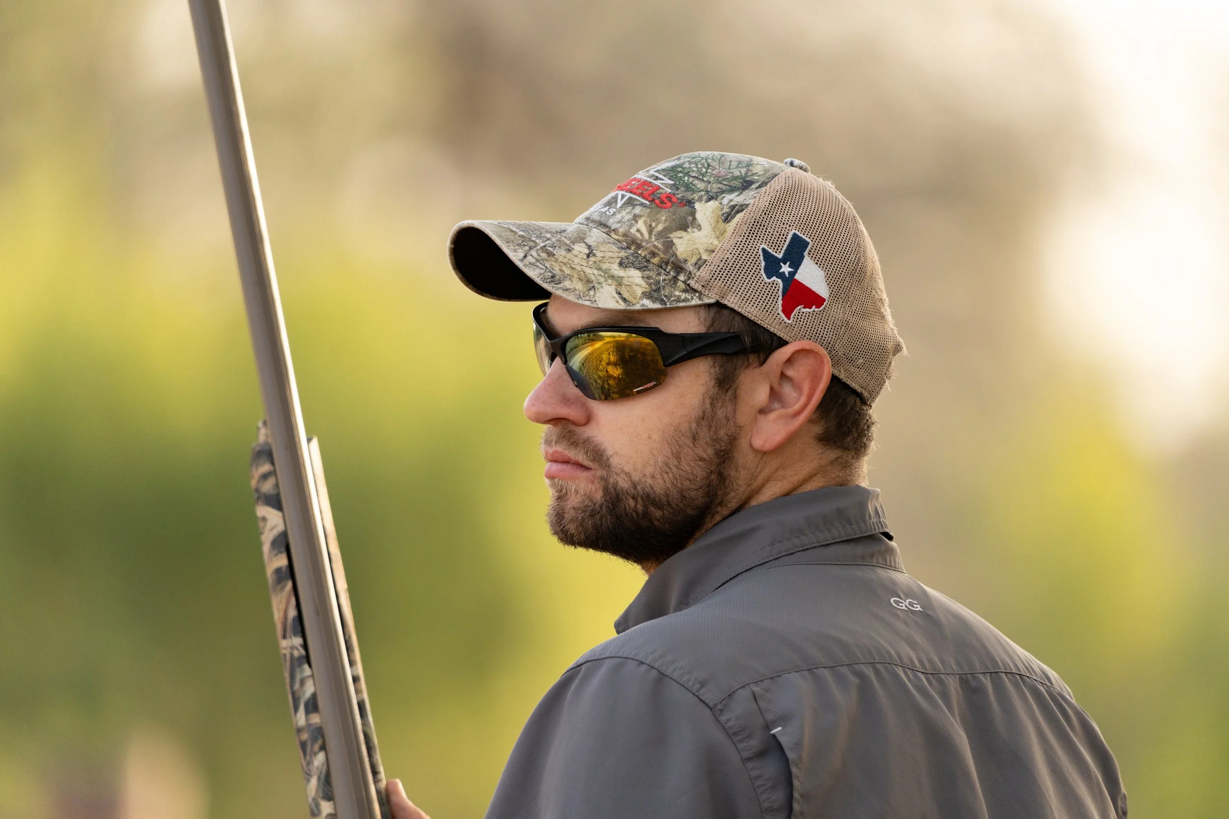 A man with a beard wearing sunglasses, a camouflage cap with Texas flag and outdoor logos, and a gray jacket, holding a rifle outdoors.