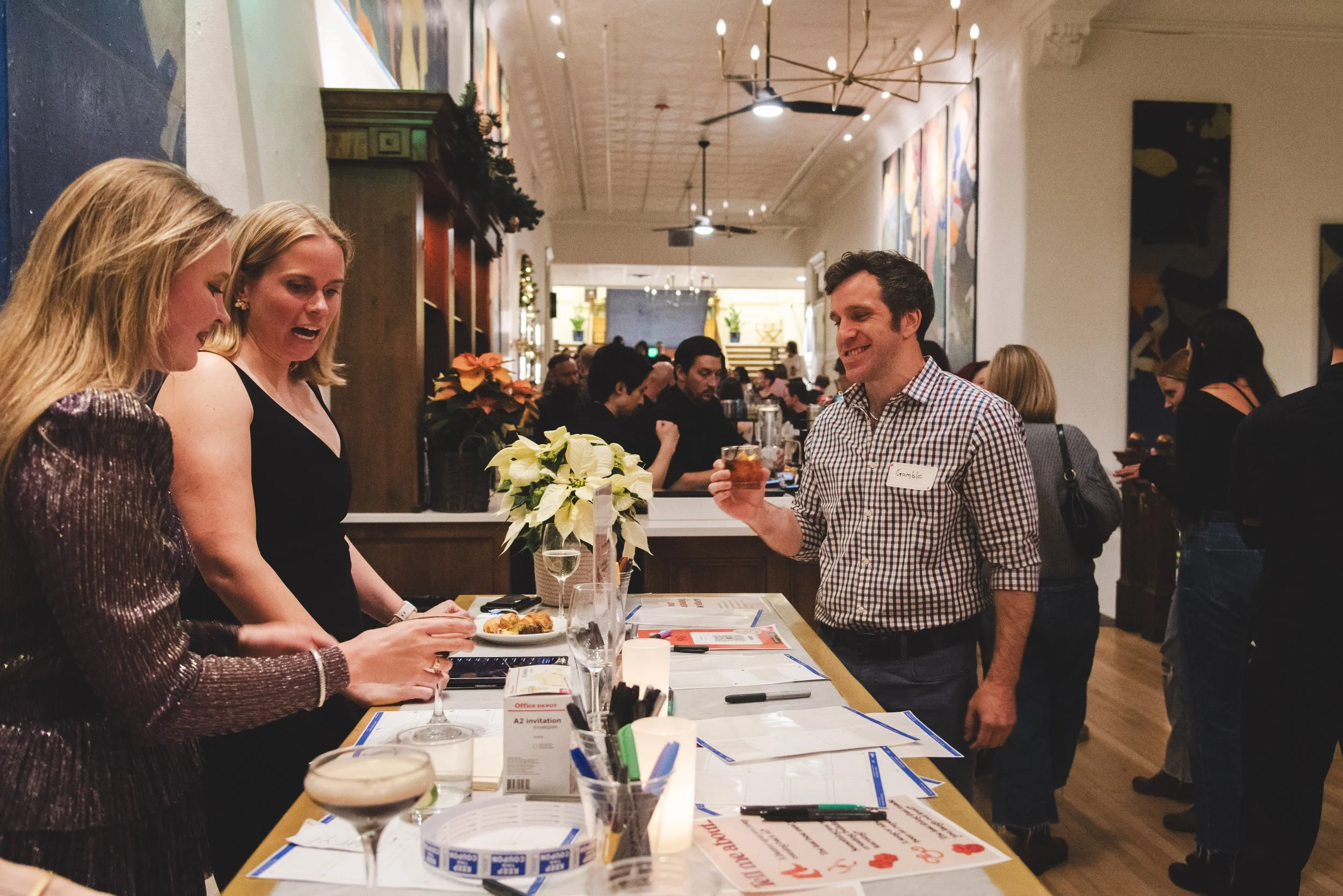 People socializing at a lively indoor event or gathering, with a table set up with drinks, food, and conference materials, in a decorated, warmly lit venue.