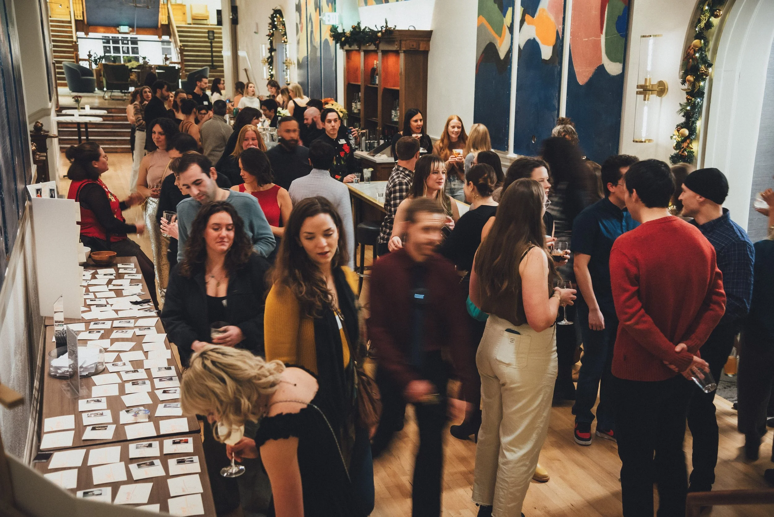 A crowded indoor holiday gathering with people mingling, some holding drinks, in a decorated room with festive garlands and artwork on the walls.