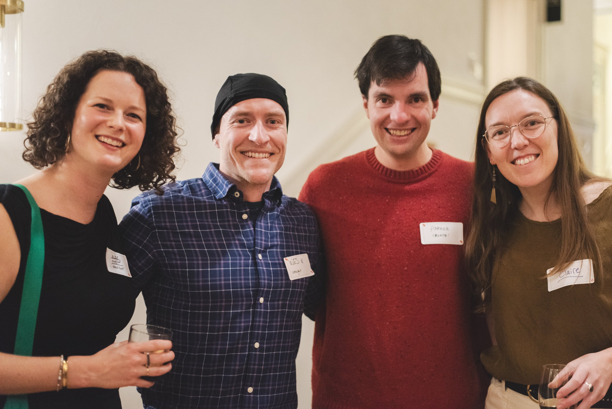 Group of four people smiling at a social gathering, one woman holding a drink, all wearing casual clothing, standing indoors with a light-colored wall in the background.