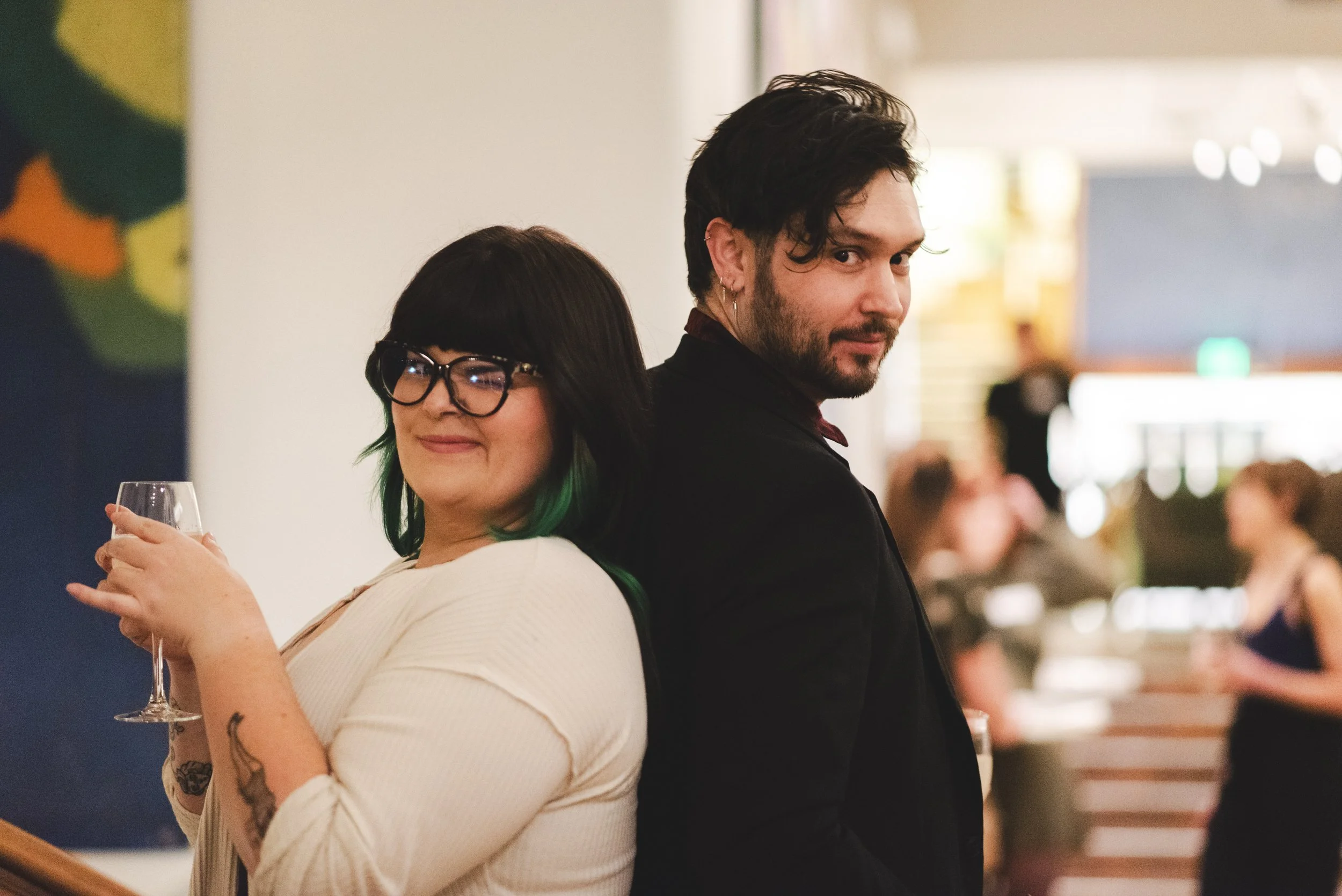A woman with glasses and green tips in her hair, holding a glass of wine, and a man with dark hair and an earring, standing back to back at Denver Dance Hall.