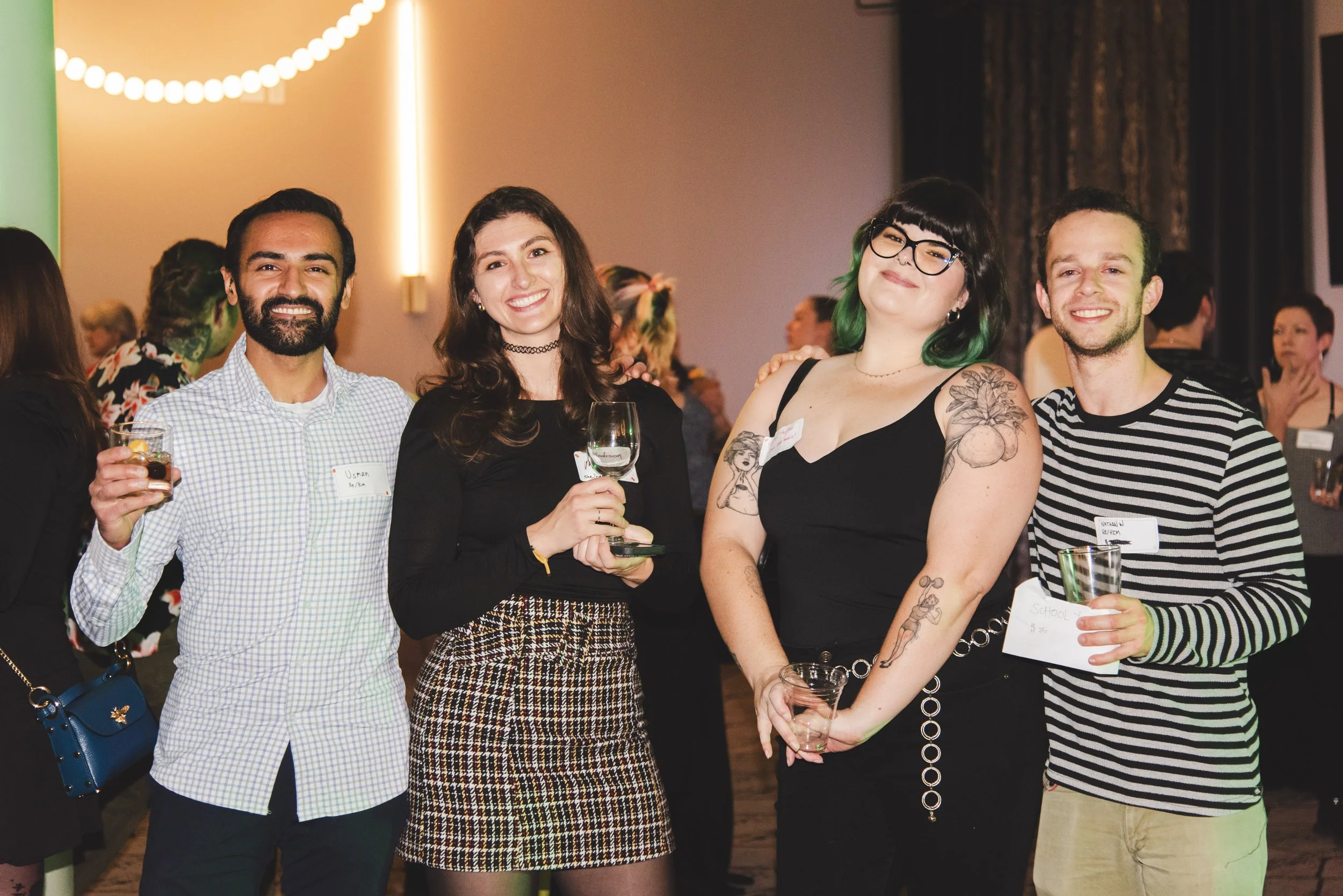Four people smiling at an indoor social event, holding drinks, with a colorful background and other attendees in the background.