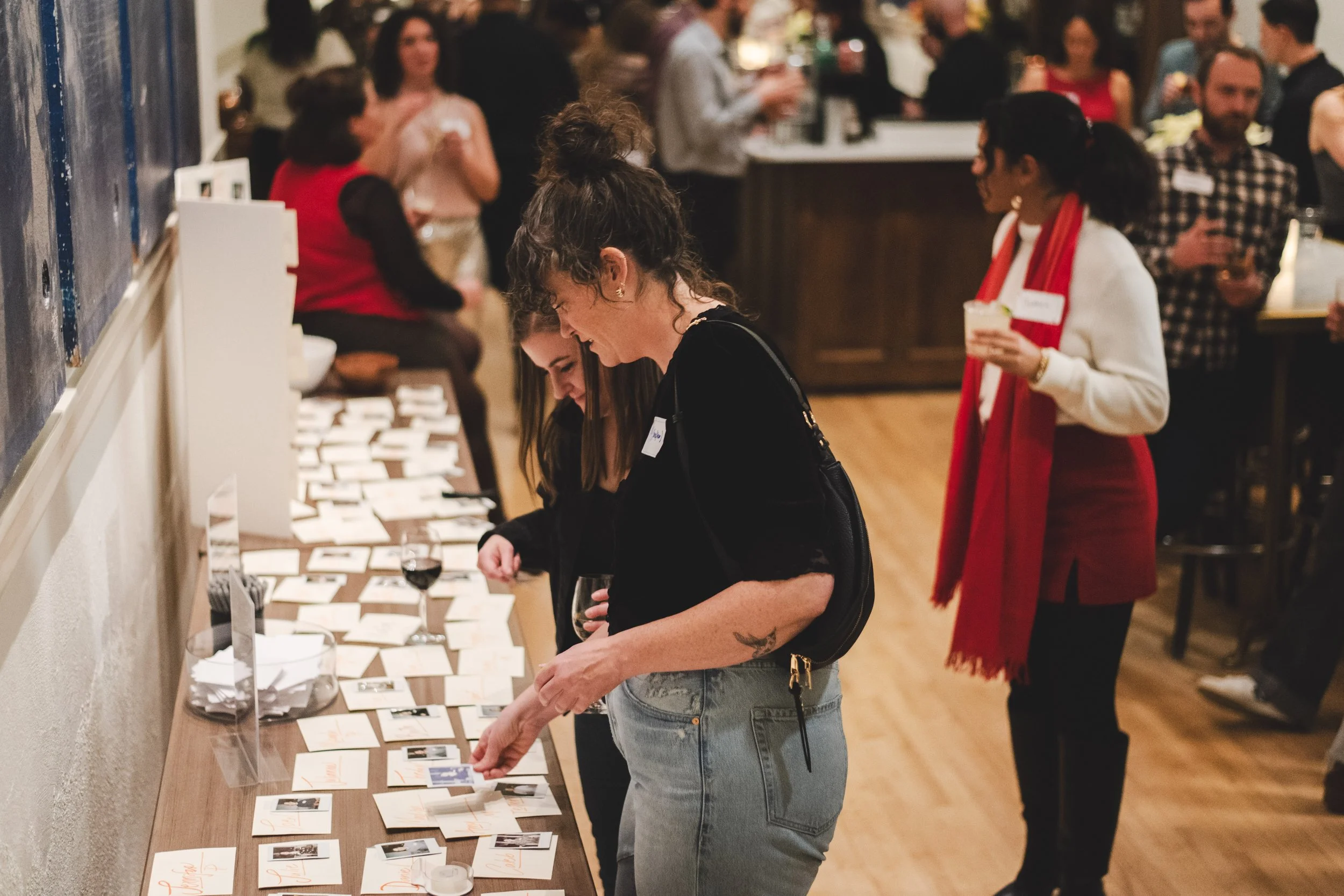 People at a social event browsing photographs and note cards displayed on a wooden table, with some holding wine glasses, in a warmly lit room with a crowded background.