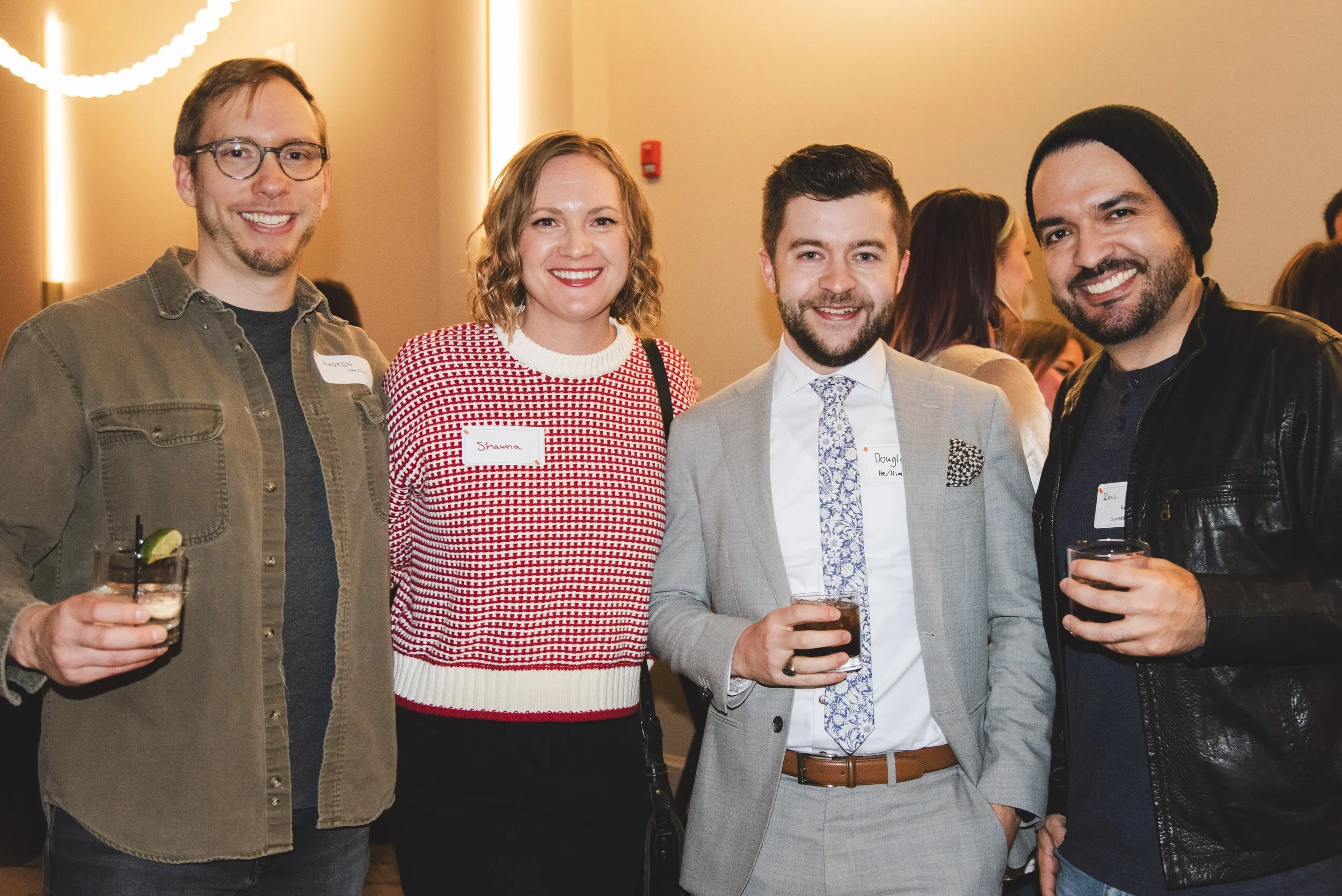 Four people smiling at a social event, holding drinks, in an indoor setting with warm lighting.