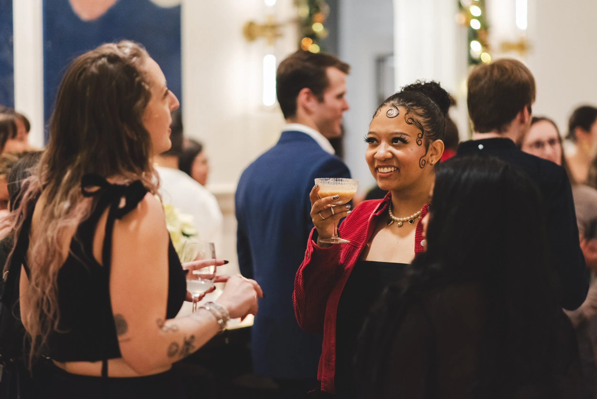 People socializing at a festive event, with women holding drinks and smiling, in a decorated indoor setting.