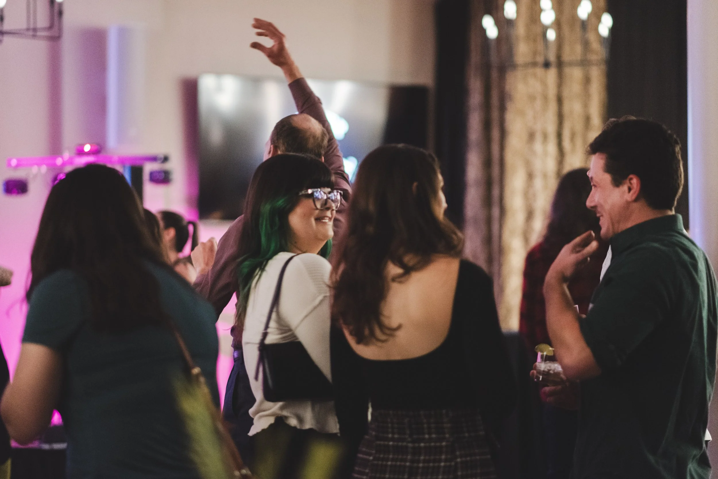 Group of people at a social gathering, engaging in conversations and smiling, with pink and purple lighting in the background.