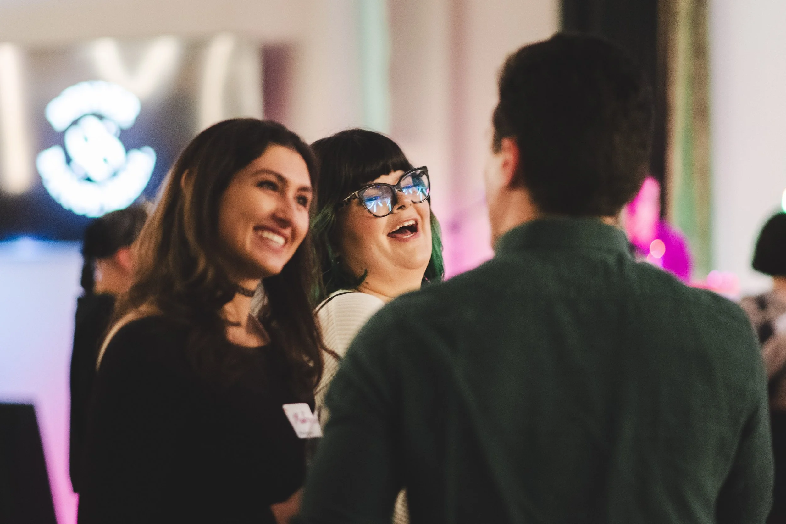 Three smiling women engaged in conversation at a social event with colorful lights in the background.