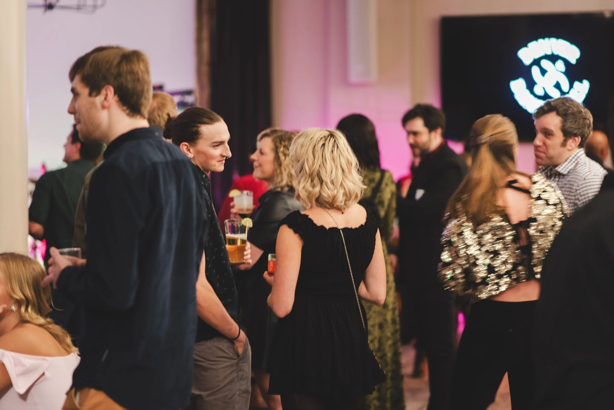People socializing at an event with a dark background and a bright logo on a screen.