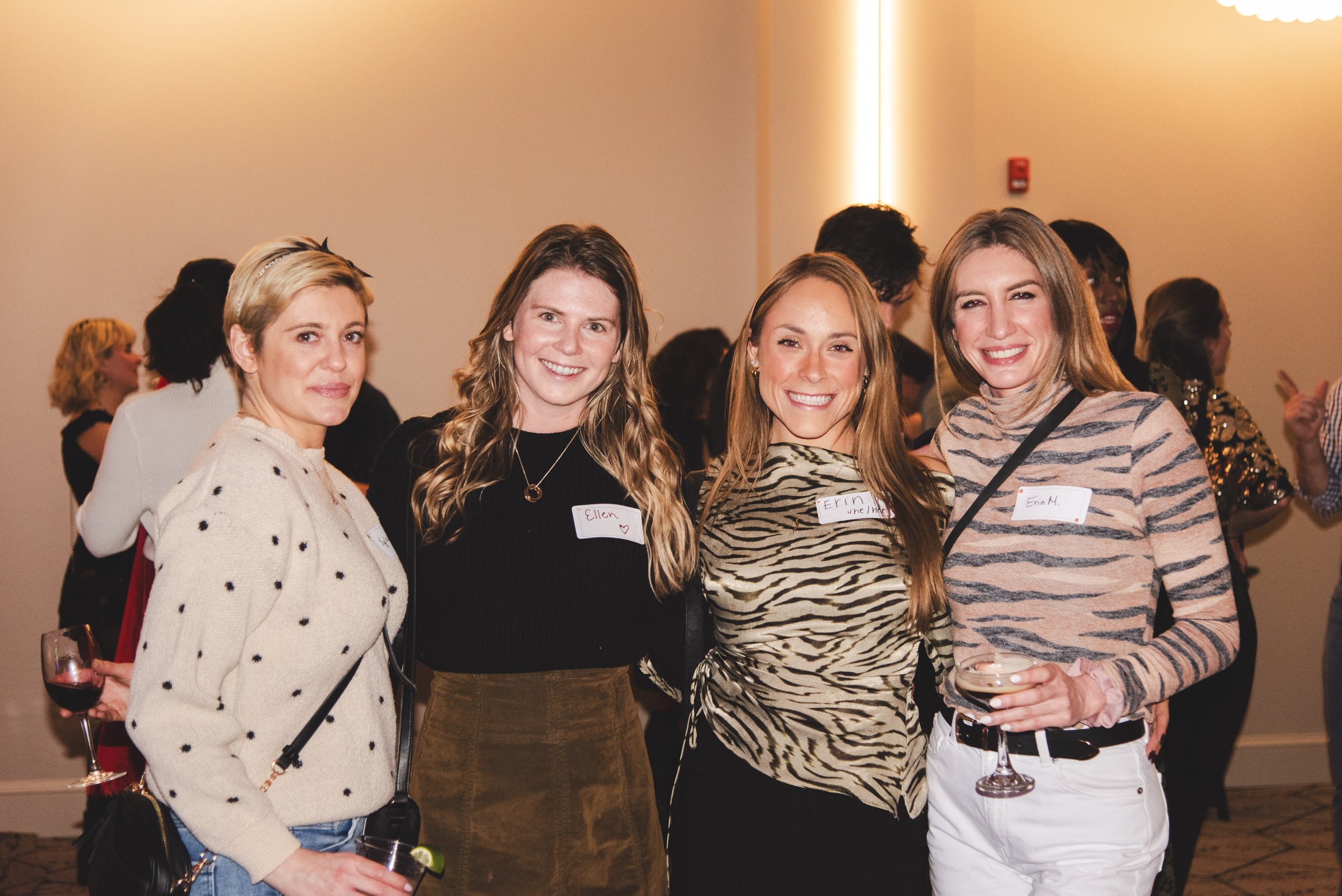 Four women standing together at a social event, smiling at the camera. They are wearing casual and stylish outfits, each with a name tag visible. One woman on the right is holding a glass of red wine, and the woman on the far right is holding a glass
