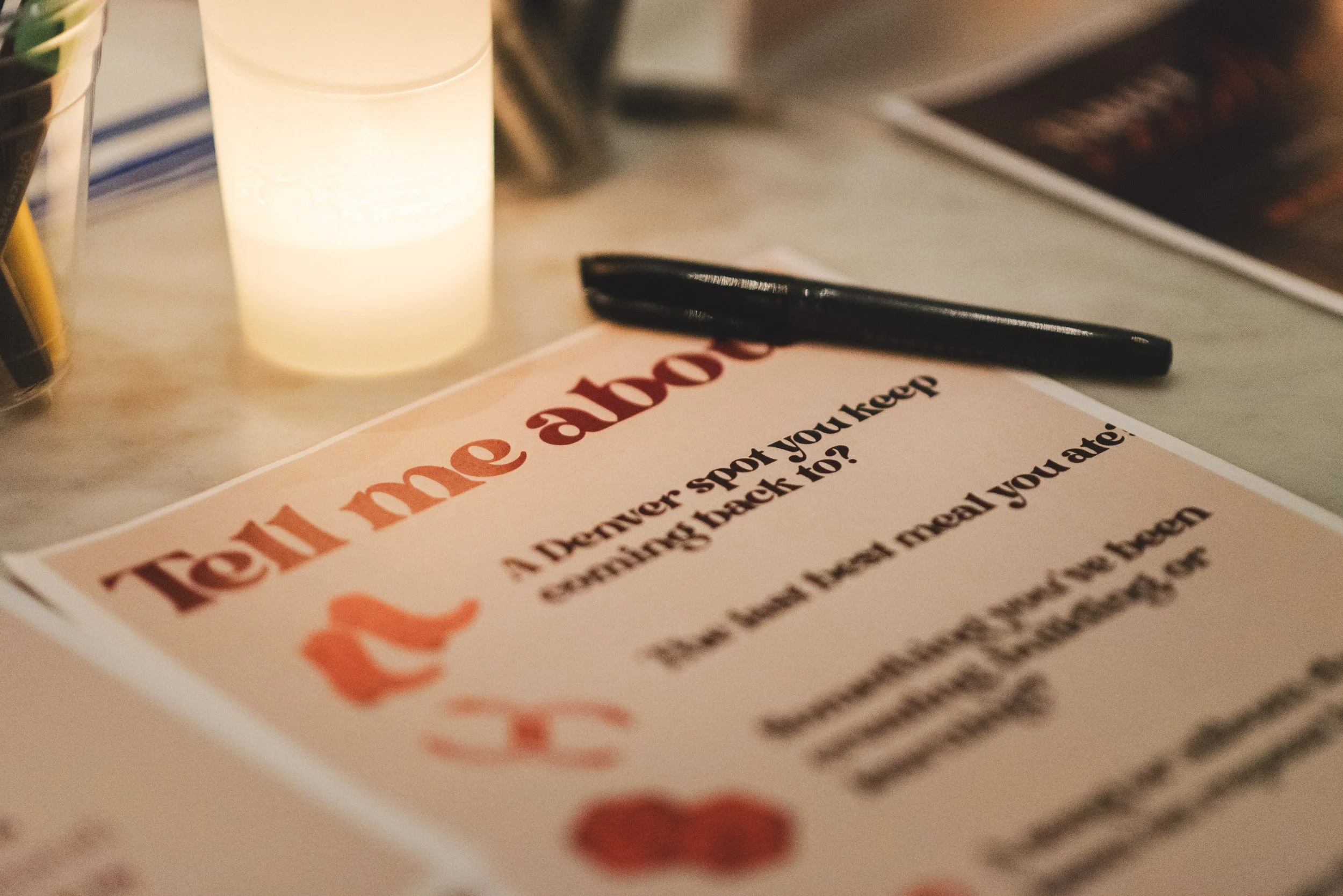 Close-up of a menu or flyer on a table, with a black pen resting on top and warm lighting highlighting the text and a lit candle nearby.