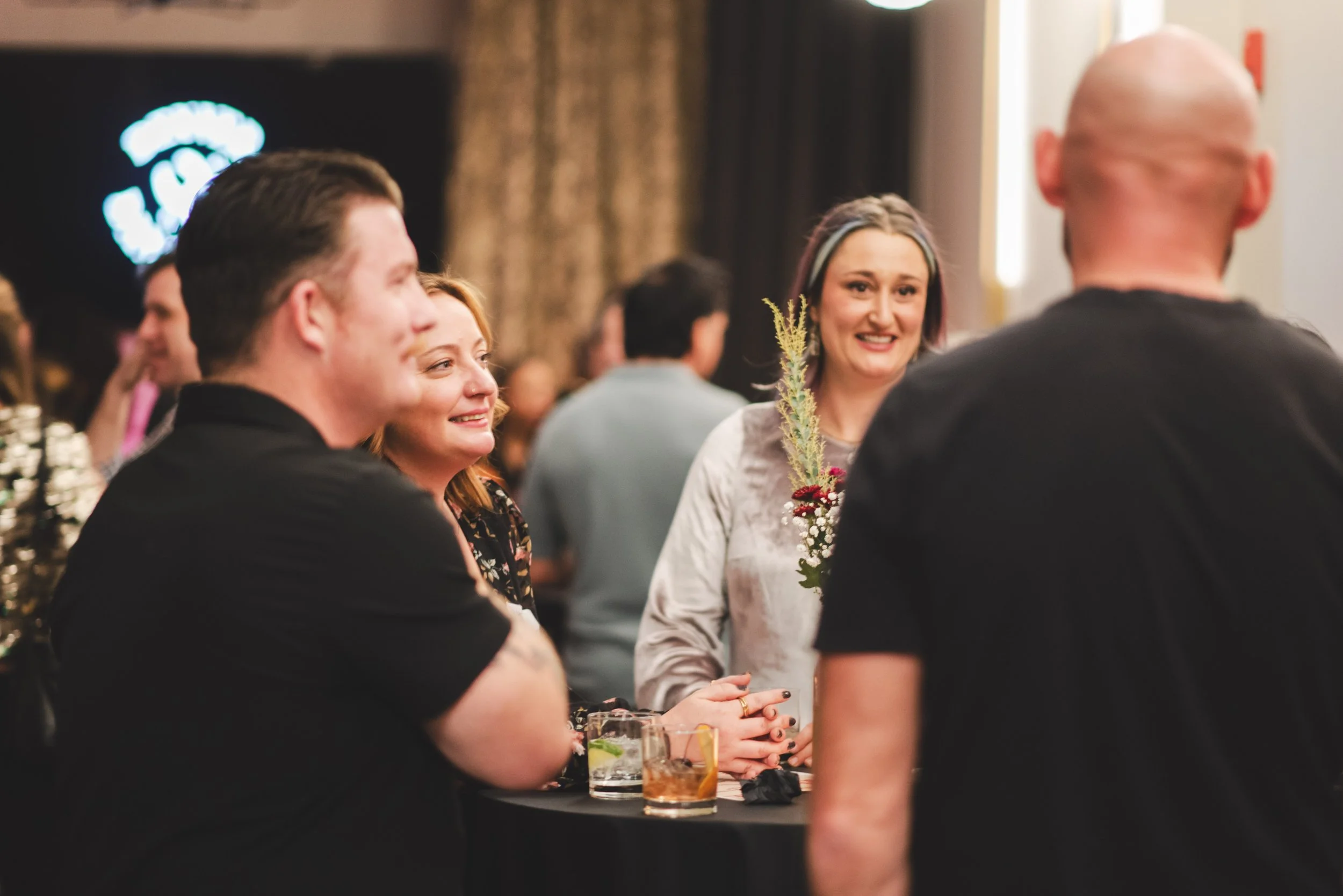 People gathered at a social event, smiling and engaging in conversation, with drinks on the table in front of them.