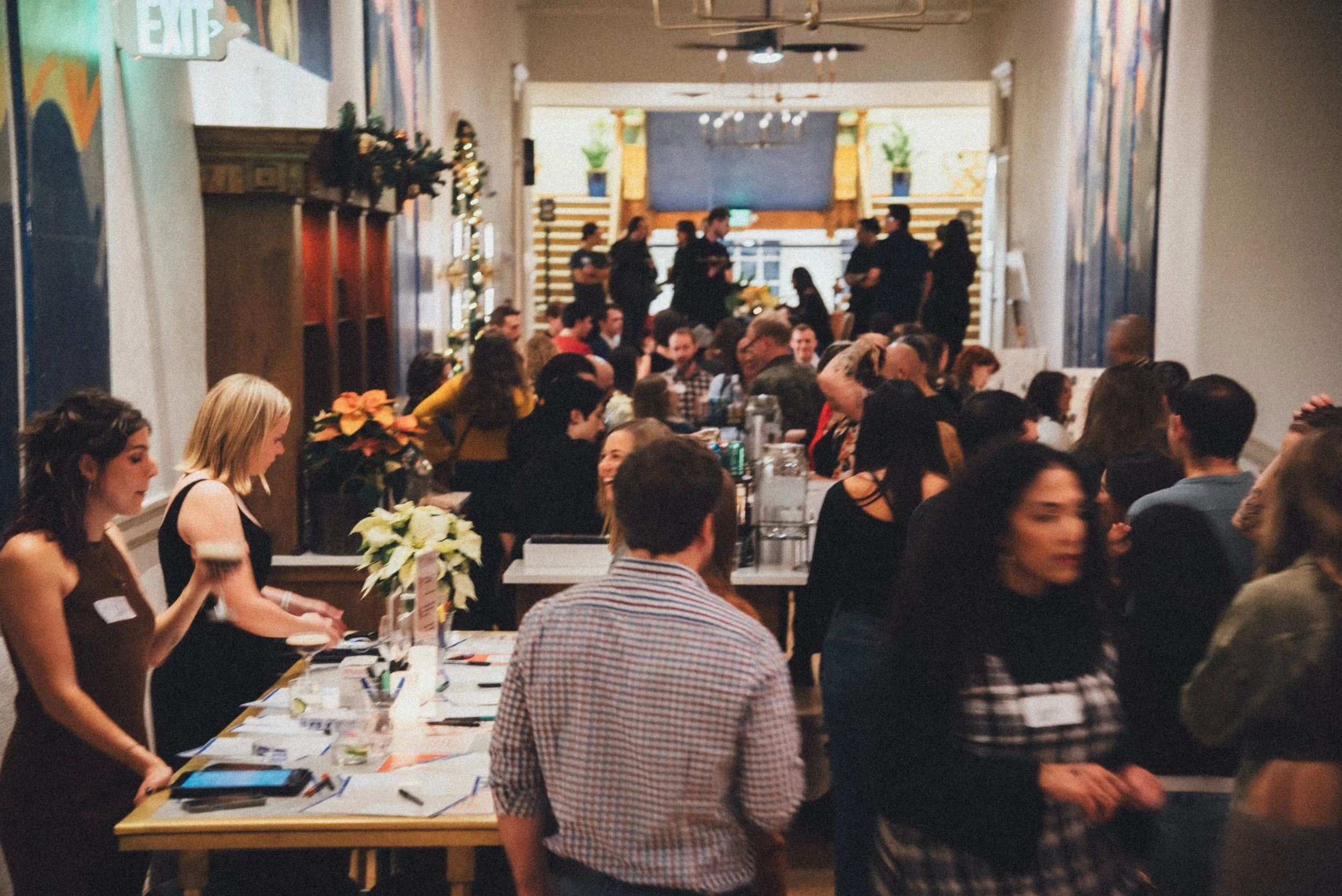 A crowded indoor event with people sitting at a long table decorated with flowers and supplies. Several people are standing or moving around, with some engaged in conversation or activities. The background shows a high ceiling, a large TV or screen, 
