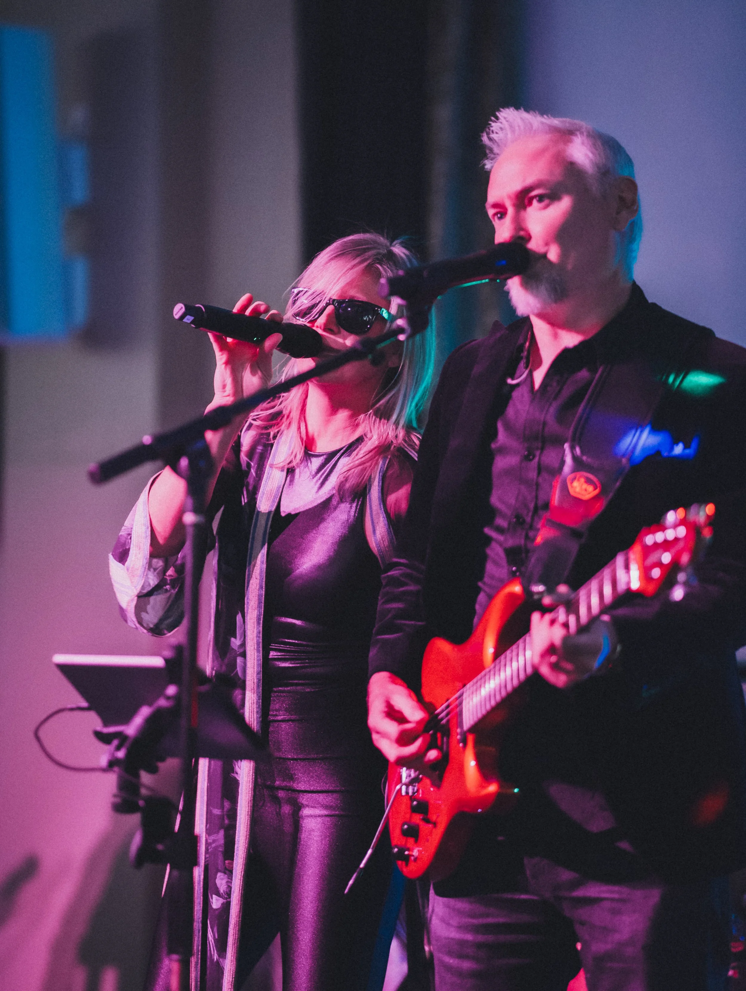 A man playing an electric guitar on stage, with a woman singing into a microphone beside him, both under colorful stage lighting.