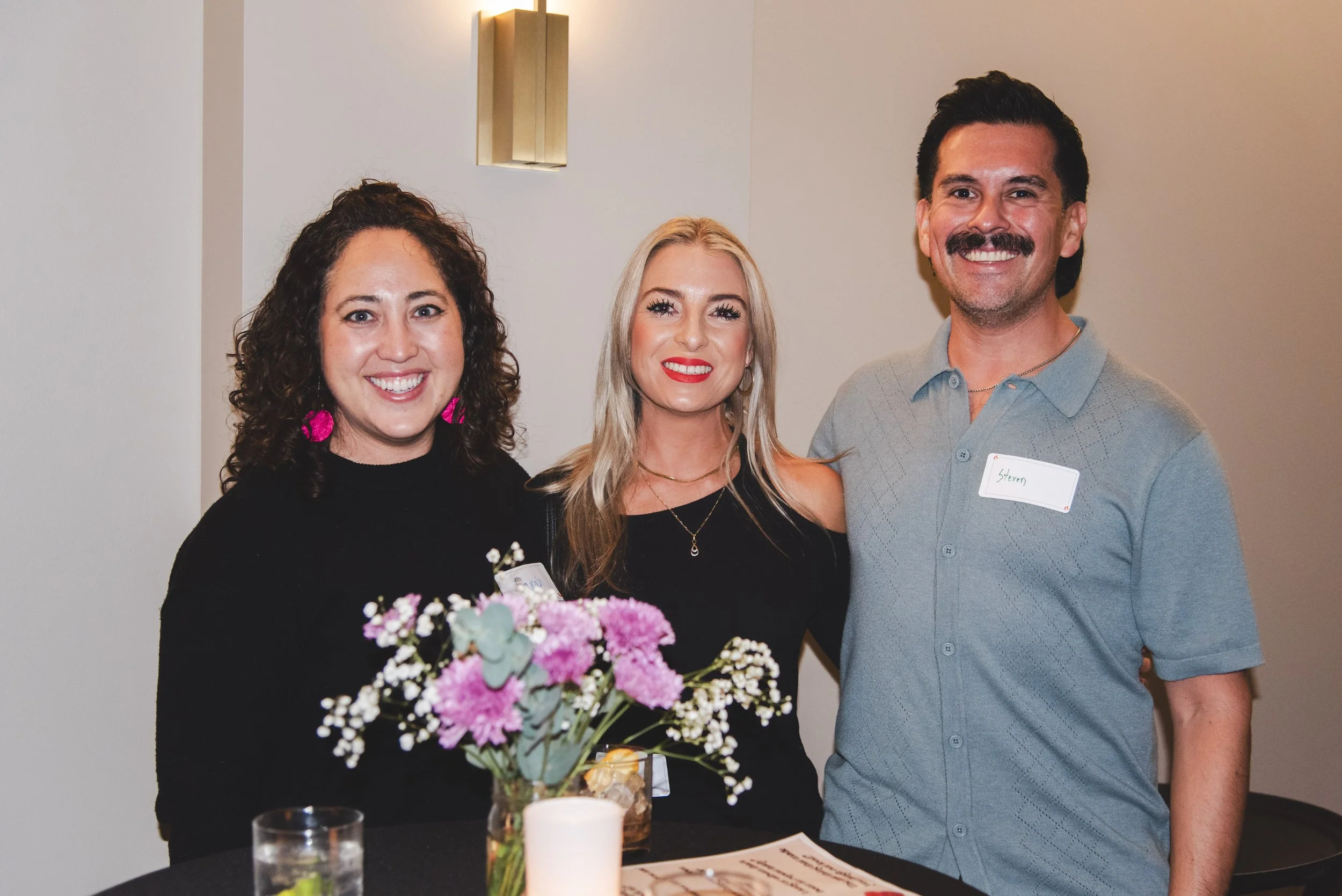 Three people smiling indoors, standing behind a table with a bouquet of pink and white flowers and a white candle. The woman on the left has curly dark hair and wears a black top with pink earrings. The woman in the middle has blonde hair, red lipsti