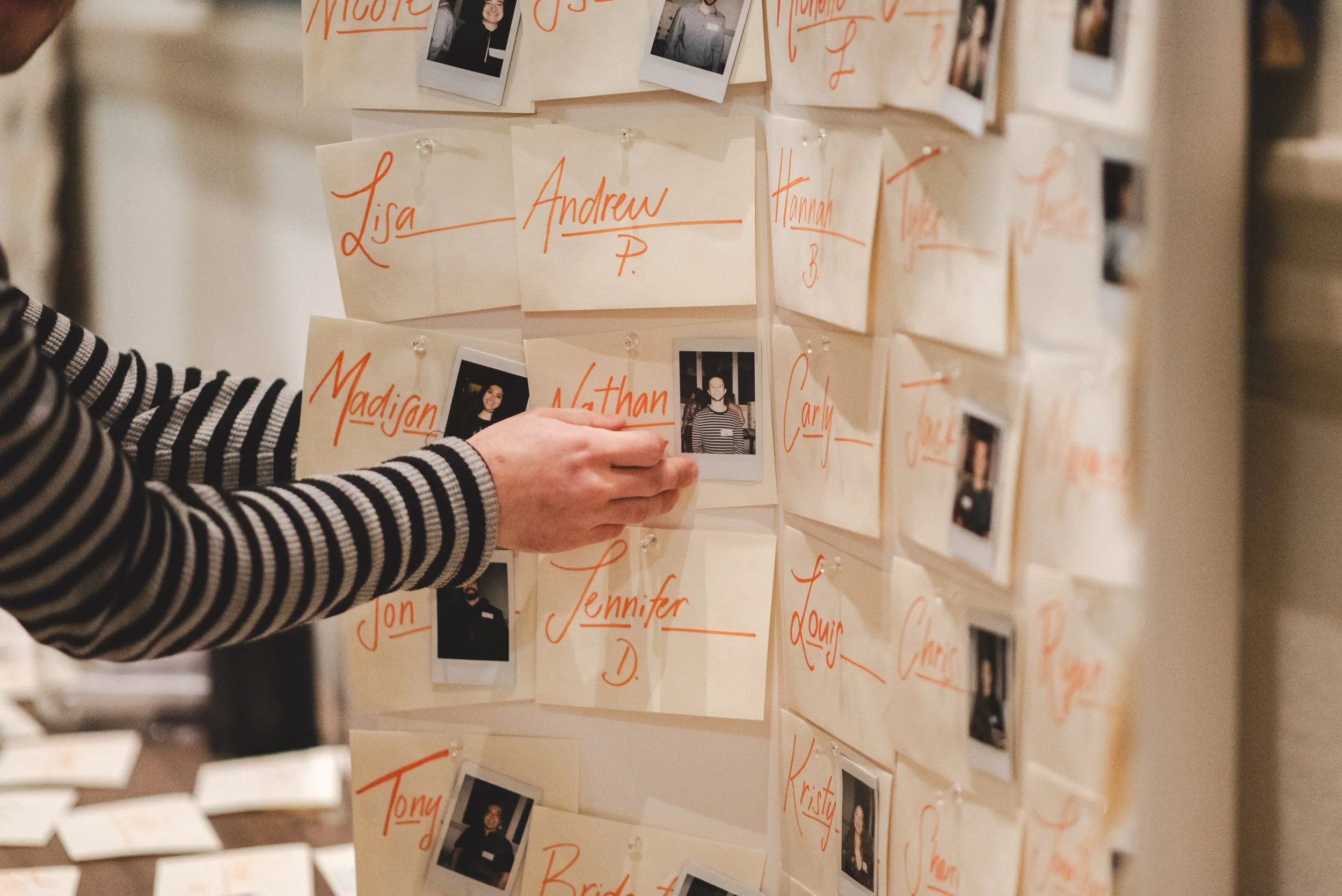 A person in a striped shirt pins a photograph on a board covered with notes, pictures, and names written in orange ink.