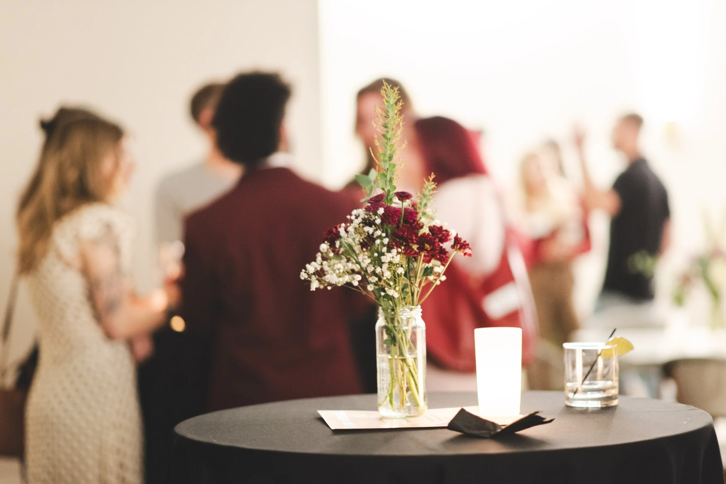 A floral centerpiece with red and white flowers in a clear glass jar on a black table in a social gathering setting with blurred people in the background.