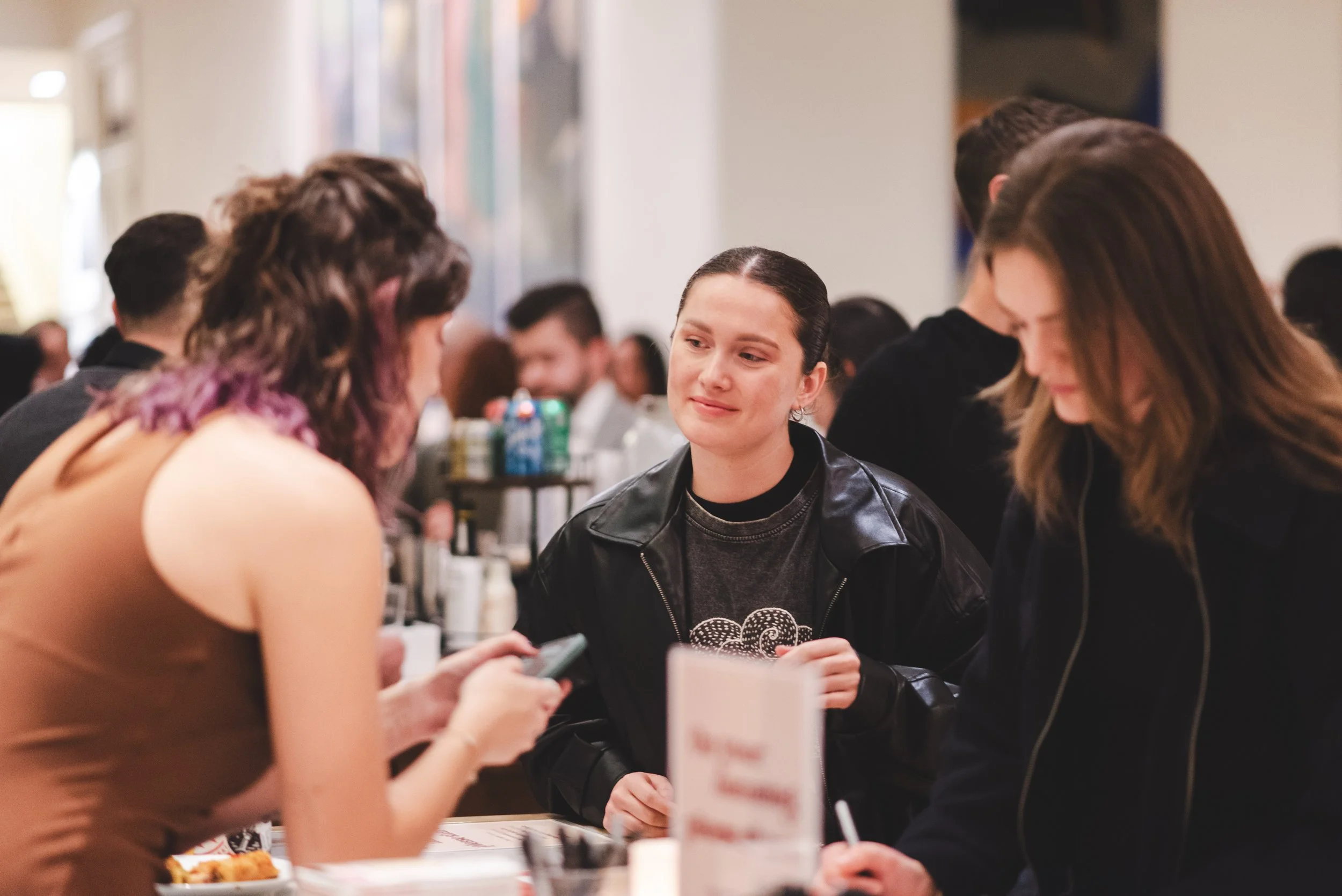 A group of people is gathered around a table at a social event, engaging in conversation and looking at a phone. The focus is on a woman with dark, slicked-back hair wearing a black leather jacket, smiling gently. The setting appears to be indoors wi