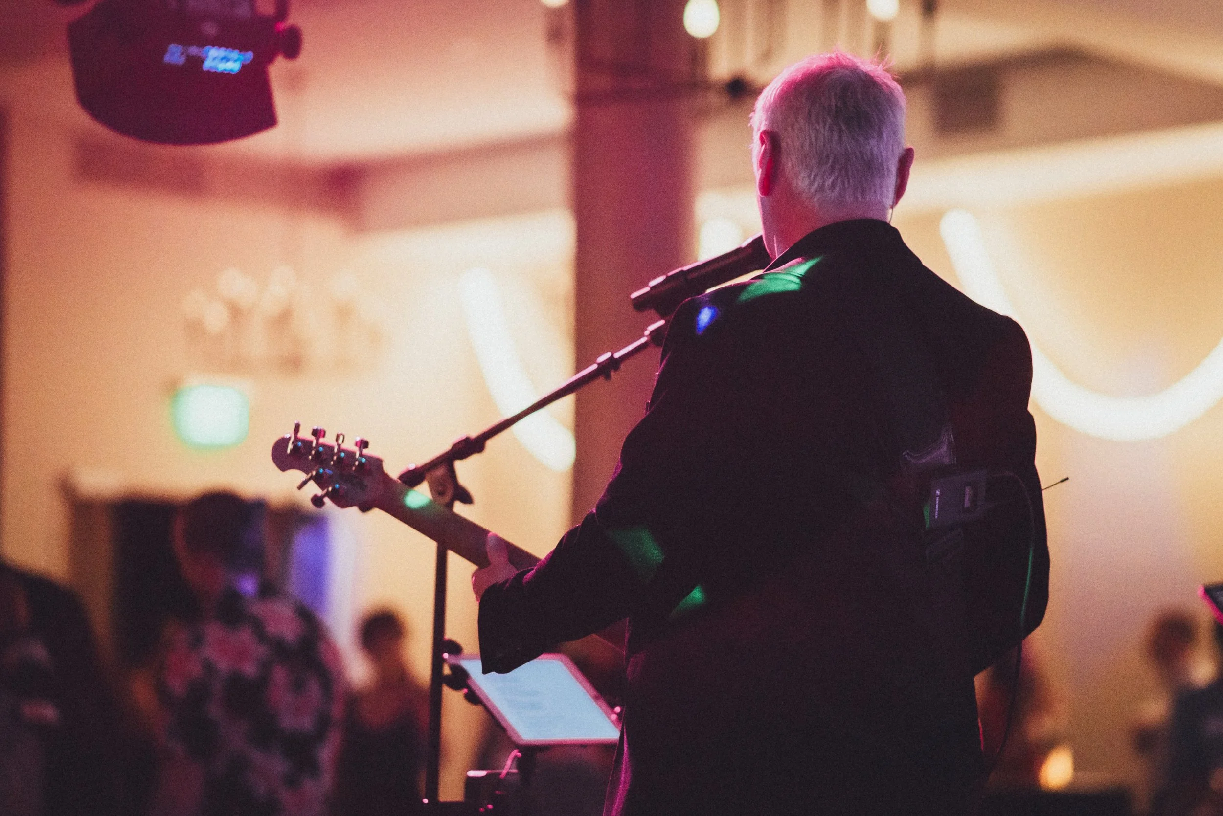 A person with short white hair, wearing a black jacket, playing an electric guitar in a dimly lit room with colorful lights, possibly at a concert or performance venue.