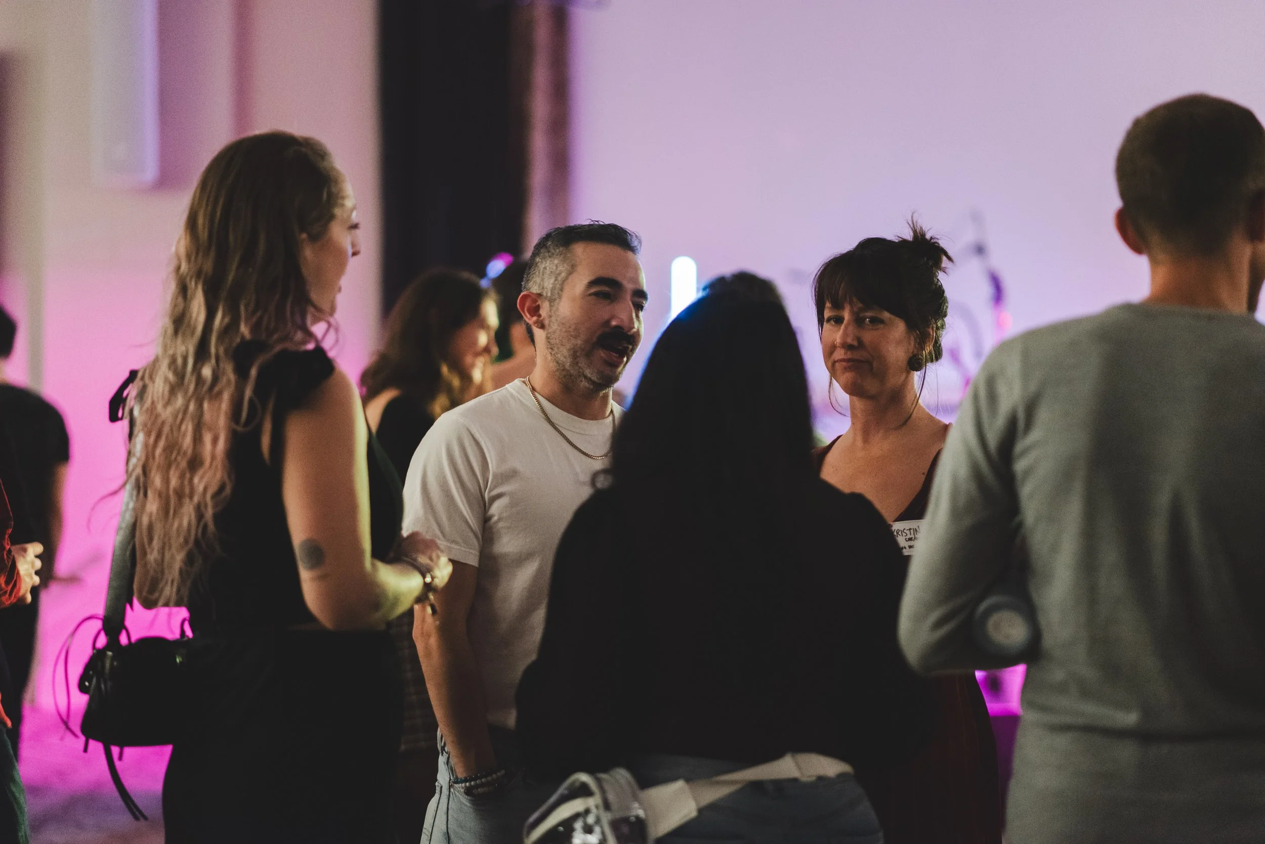Group of people engaging in conversation indoors, with colorful lighting in the background.