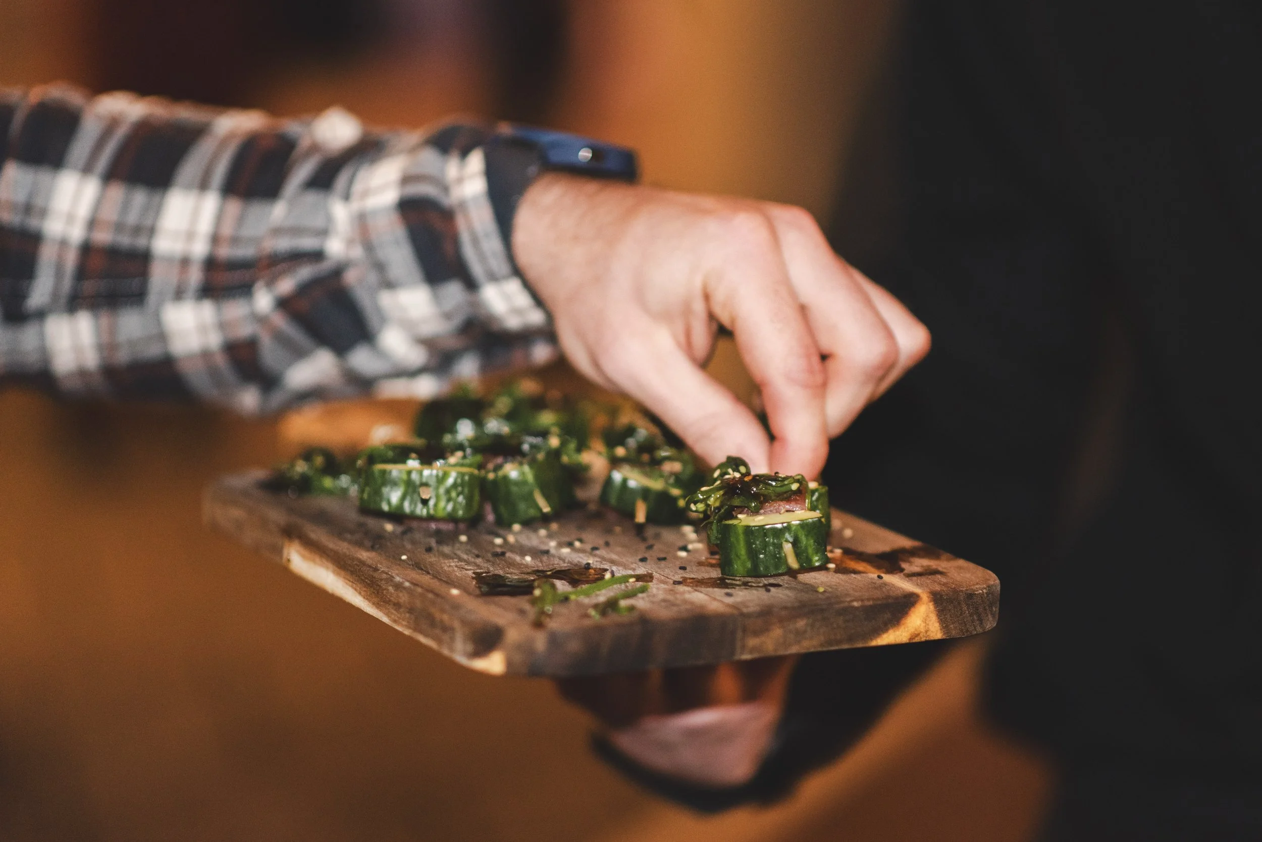 A person in a plaid shirt is preparing cucumber sushi rolls on a wooden cutting board.