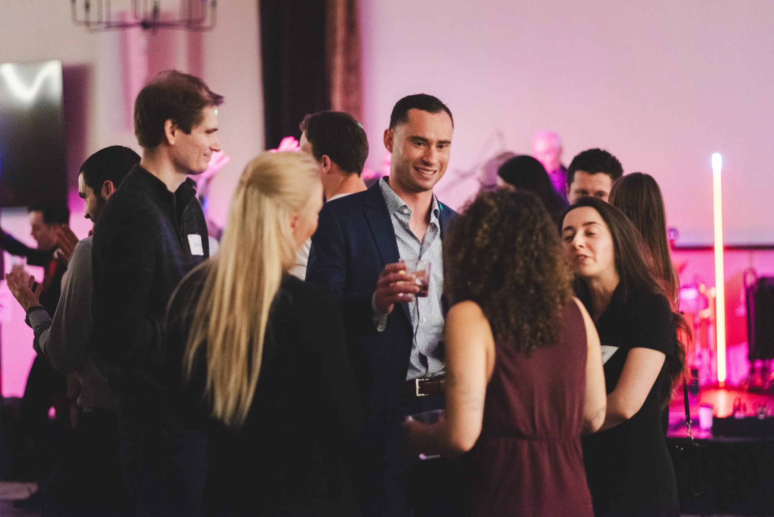 People socializing at a party or event in a room with pink and purple lighting, engaging in conversation and holding drinks.