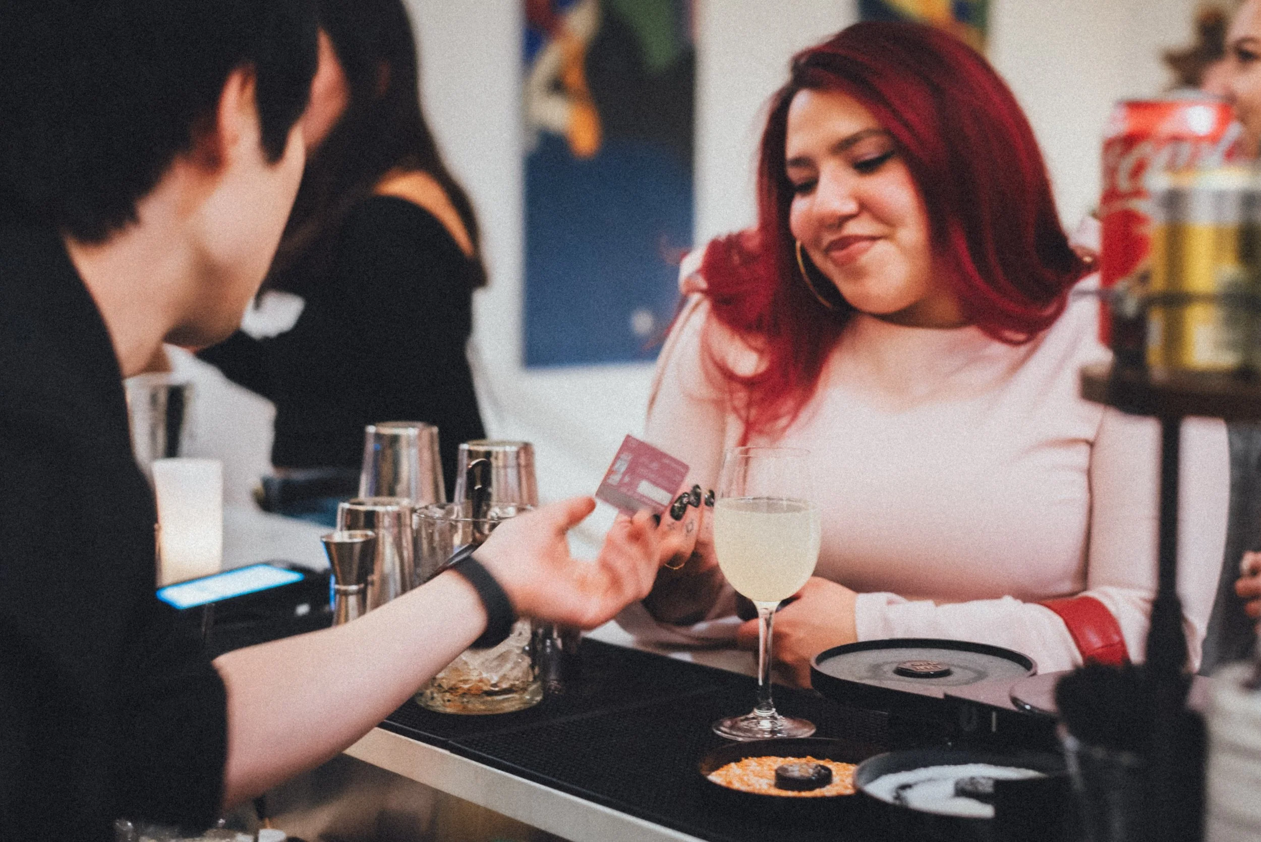 A woman with red hair wearing a light pink top in a bar or restaurant, receiving a cocktail drink from another person. There are glasses and plates on the counter, and a person in the background. The woman has a slight smile and appears to be enjoyin