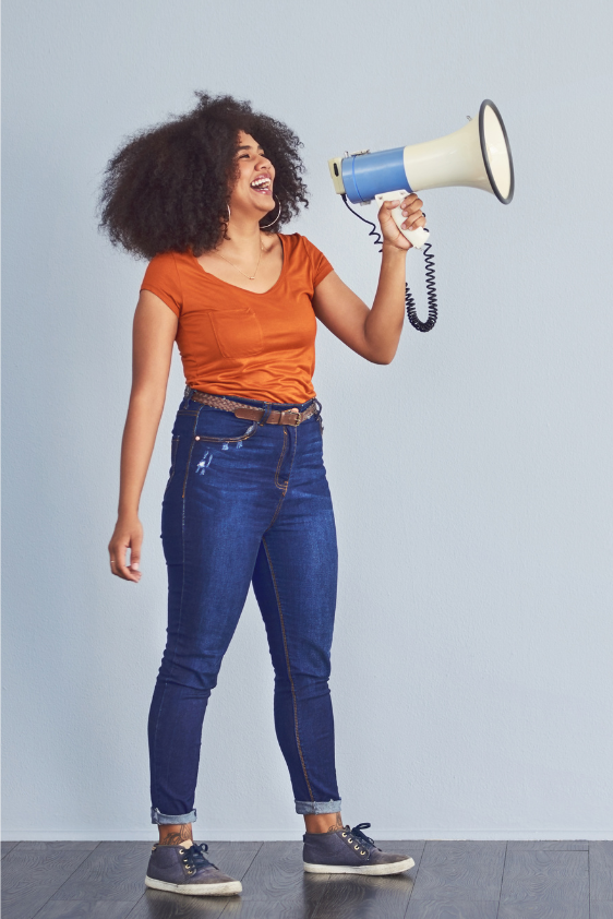 A woman with curly hair wearing an orange t-shirt and jeans, holding a megaphone and smiling.