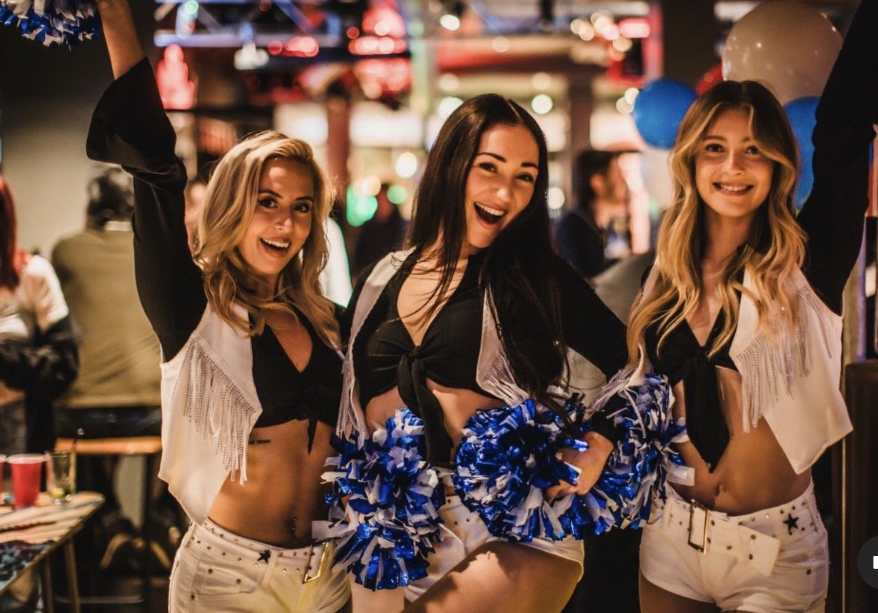 Three smiling cheerleaders in western-style fringe jackets holding blue and white pom-poms, entertaining guests at a lively bar event and bringing energy to brand activations and watch parties.