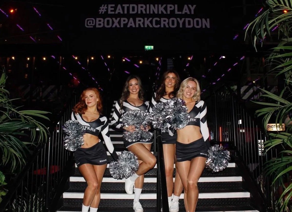 Cheerleaders in black and white uniforms with silver pom-poms posing on the stairs at Boxpark Croydon, creating a lively atmosphere for venue events, sports nights and brand promotions.