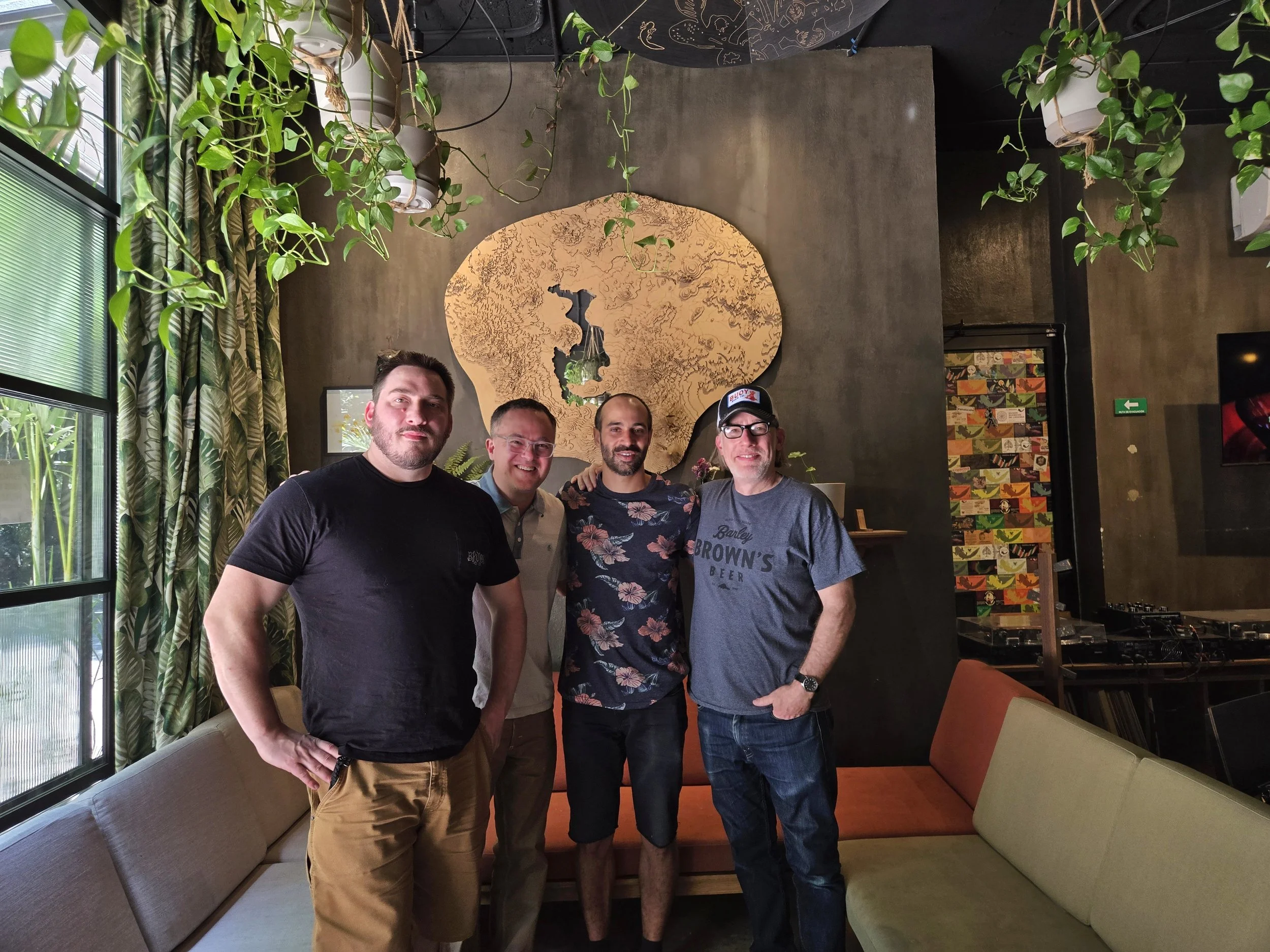 Four men standing together in a café or lounge, smiling for the camera. The background features a wooden art wall, greenery, and colorful patterned decor.