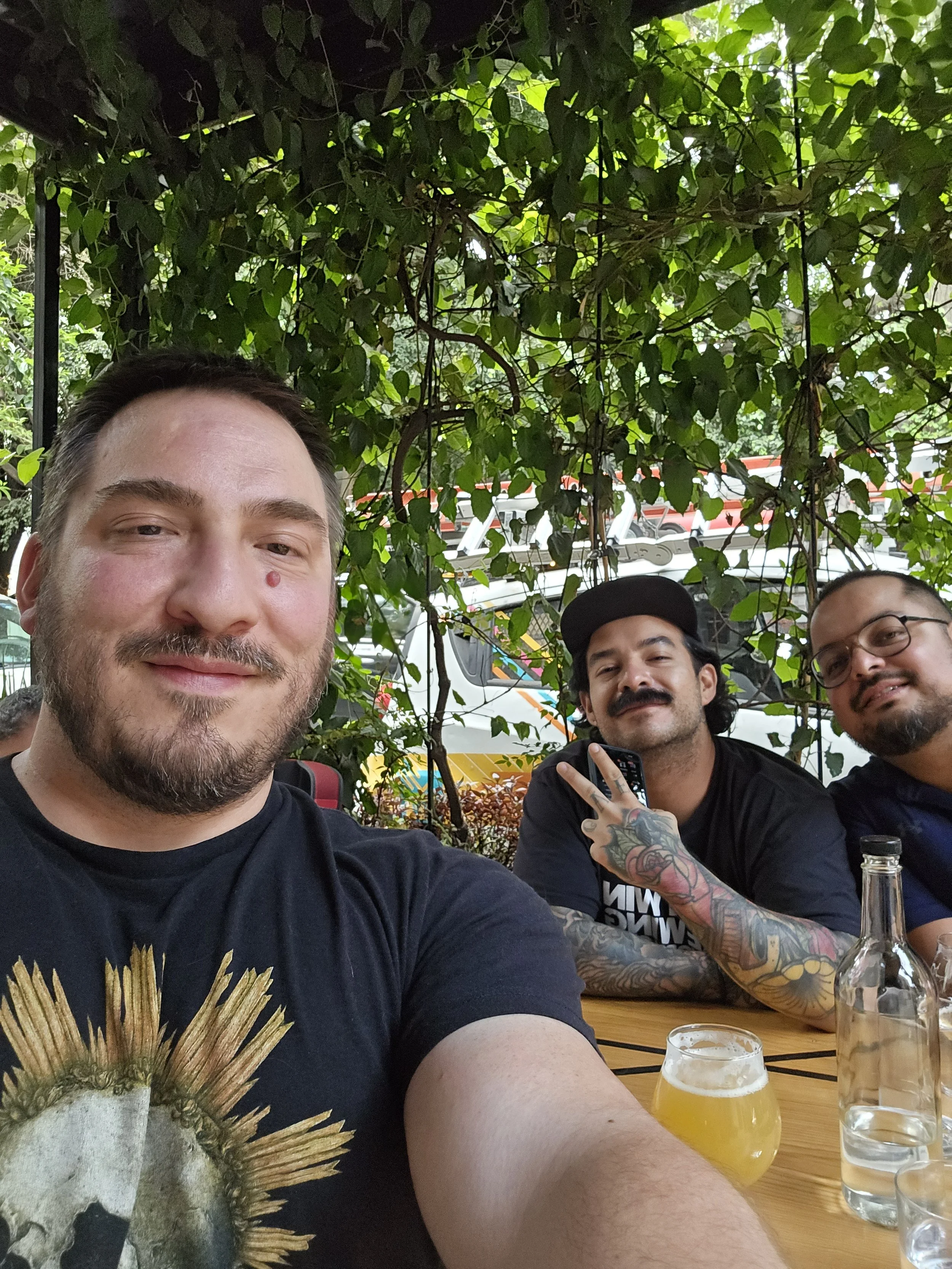 Three men sitting at a table outdoors under green leafy vines, smiling at the camera. The table has a glass of beer, a bottle, and a glass of water.