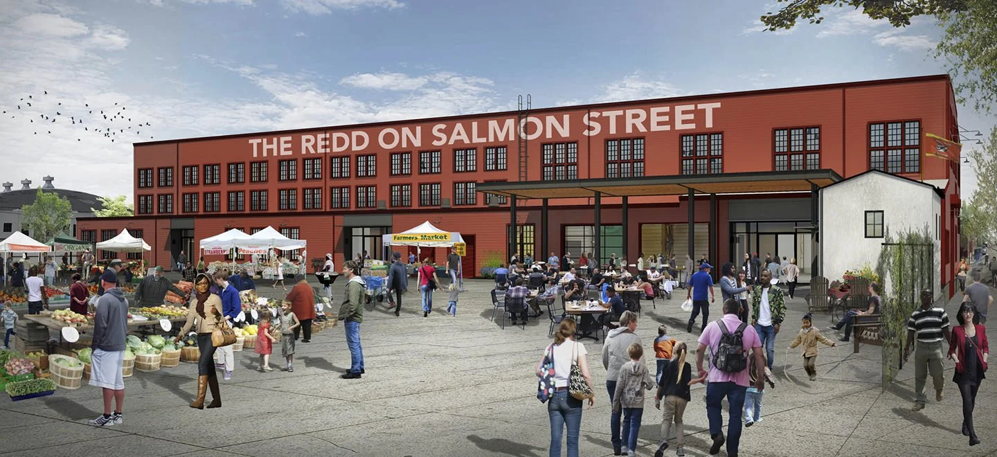 A busy outdoor marketplace in front of a large red building with a sign that reads 'The Redd on Salmon Street.' People are shopping at vendor booths, sitting at outdoor tables, and walking around in the open area.