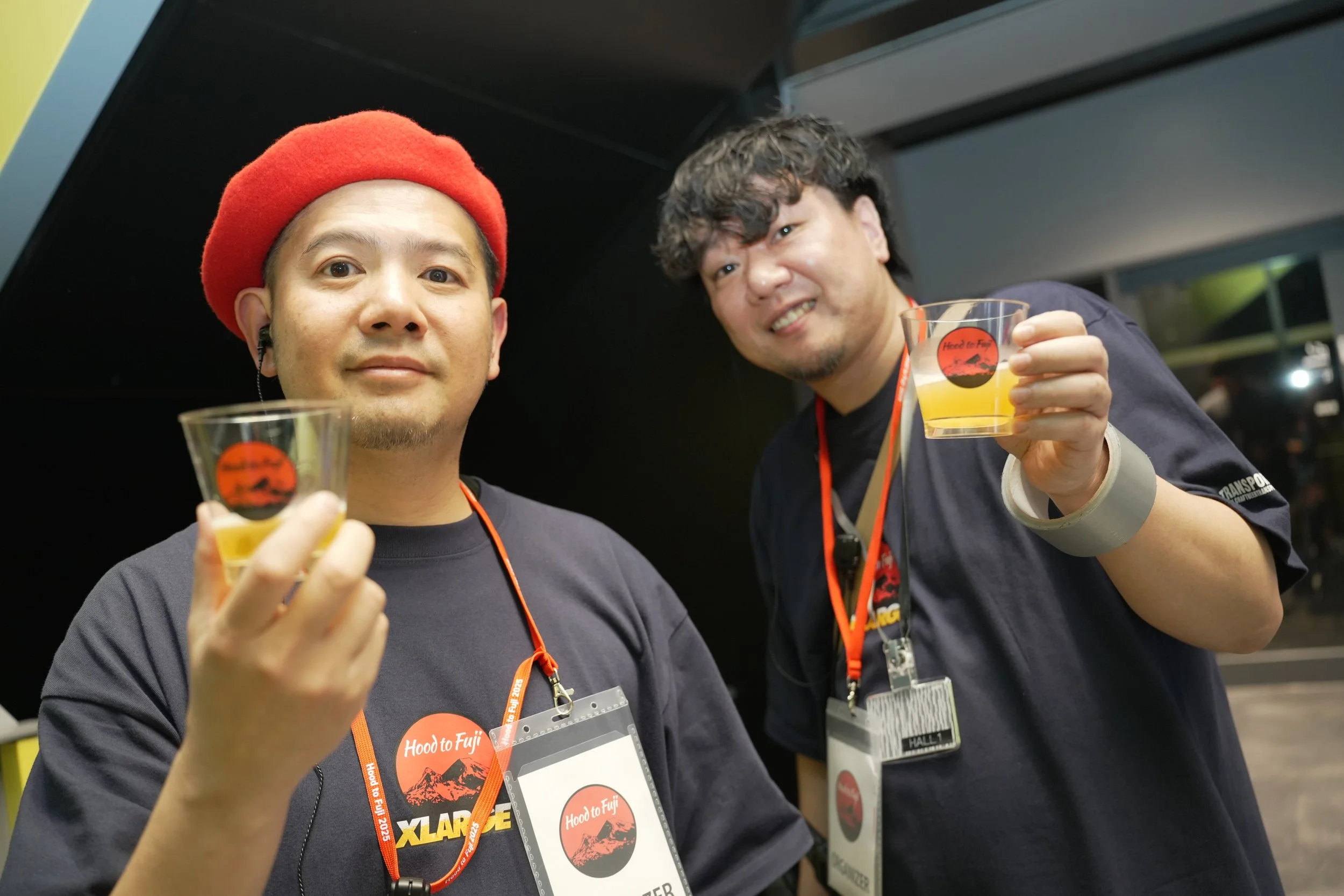 Two men at an event holding drinks in clear glasses with a logo, wearing event badges and lanyards, smiling at the camera.