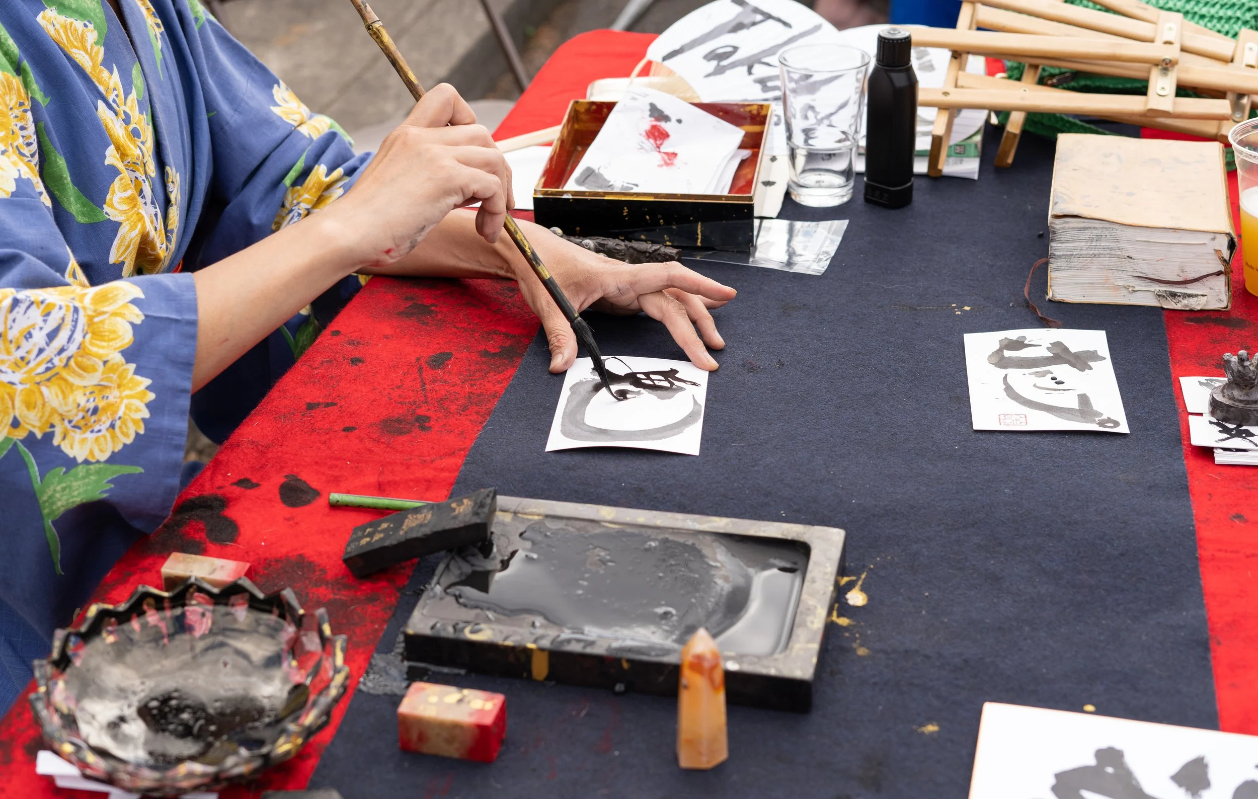 Person practicing Japanese calligraphy with black ink and brush on paper, surrounded by art supplies on a table covered with a red and black cloth.