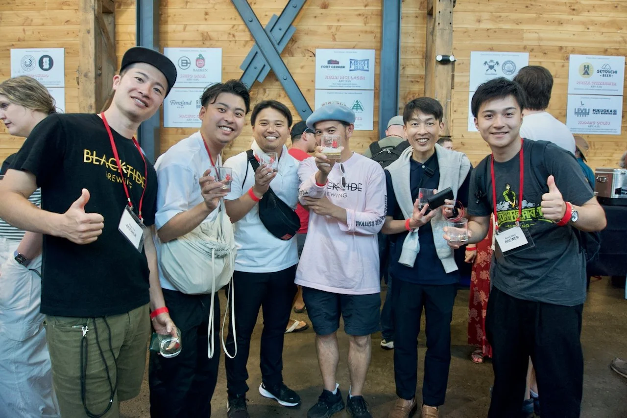 Group of six young men at a beer tasting event, holding glasses, smiling, in a wooden hall.