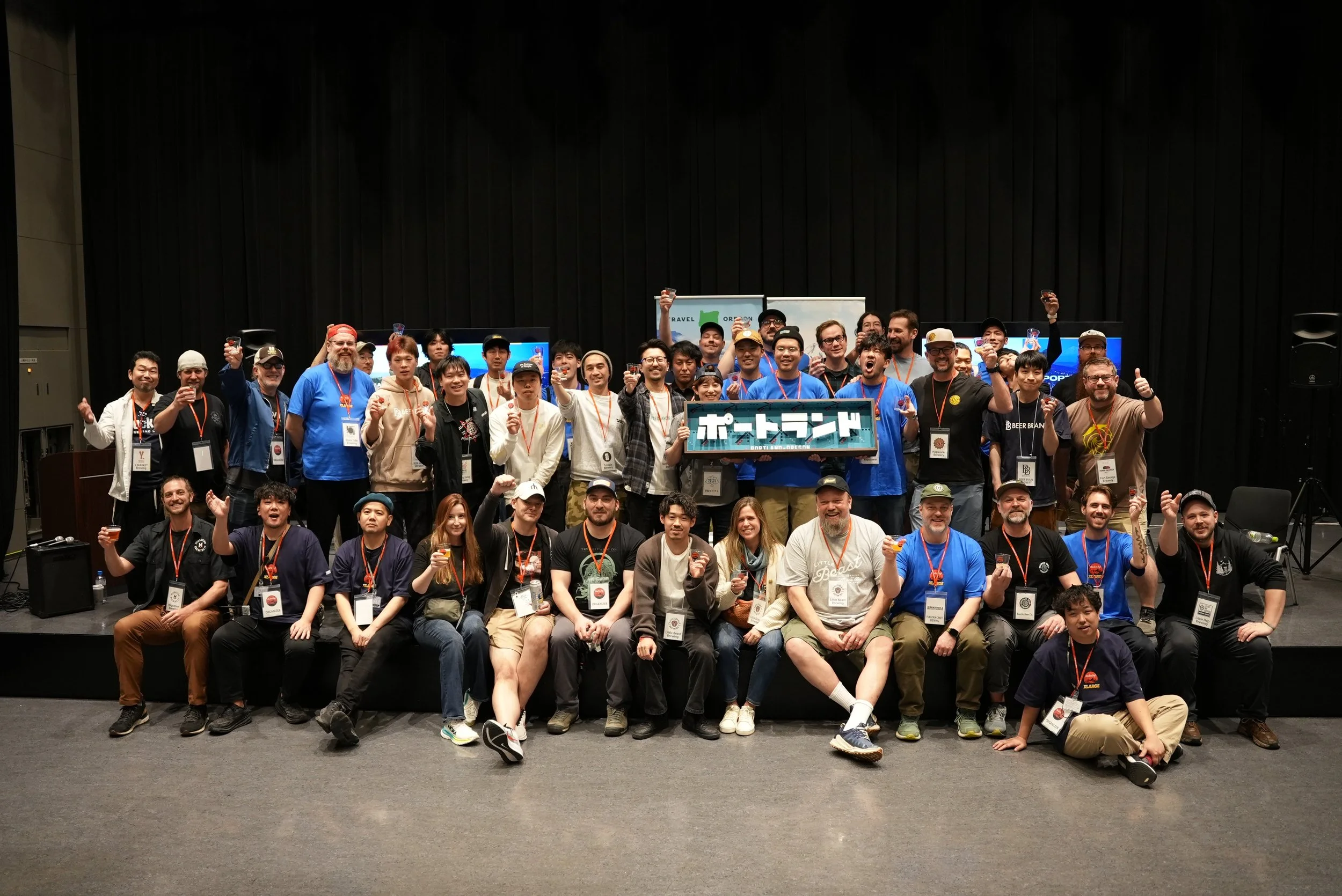 Group of diverse people on stage celebrating, some holding drinks, with a sign in Japanese, at an event in an auditorium.