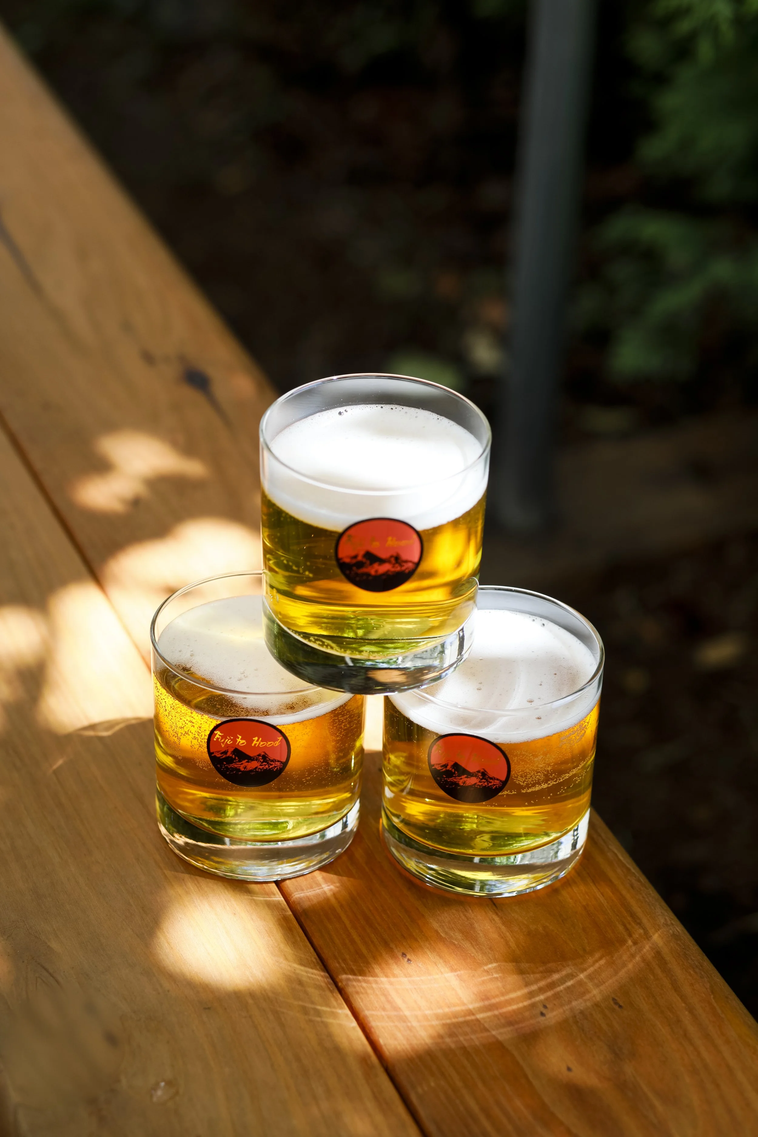 Three glasses of beer with foam, one stacked on top of two on a wooden surface with sunlight and shadows.